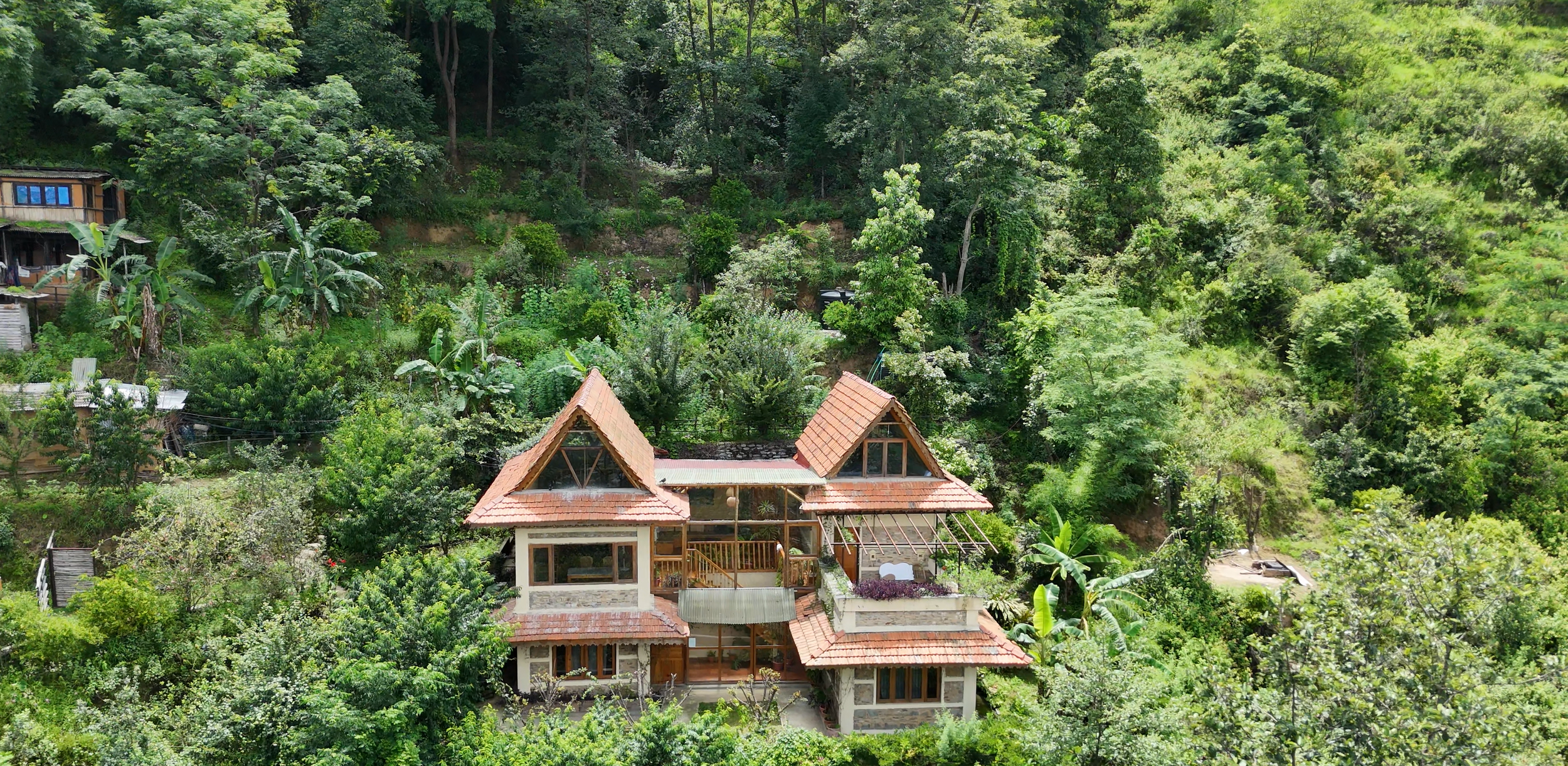 A house with a red-tiled roof, surrounded by dense green forest and trees, in a hilly area.