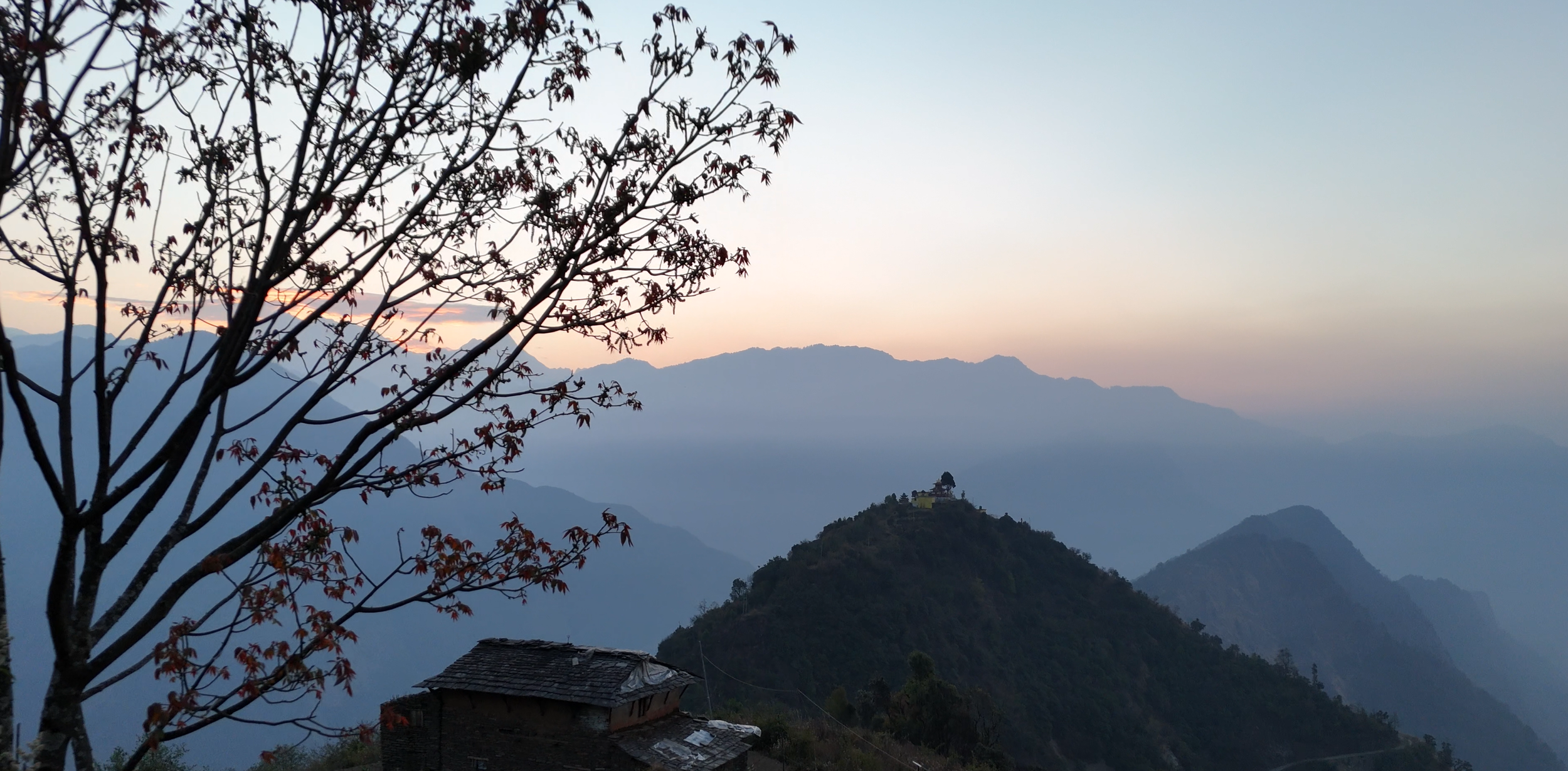 Mountain landscape during sunrise with layers of mountains, a hill with a building and trees in the foreground, and a tree with red leaves on the left.