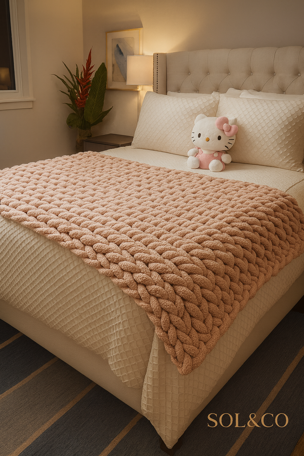 A cozy bedroom with a beige, tufted upholstered headboard, cream-colored bedding, and a pink chunky knit blanket. A Hello Kitty plush toy sits on the bed. To the left, a nightstand with a lamp, a framed artwork, and a potted plant with large green le