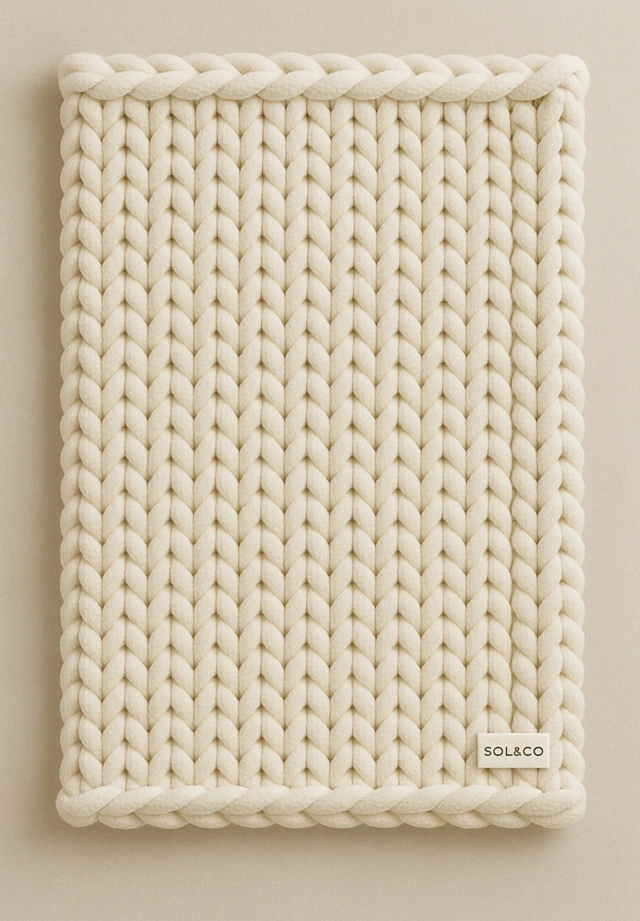 A cream-colored chunky knit blanket with a braided border and a small label reading "SOL&CO." in the bottom right corner.
