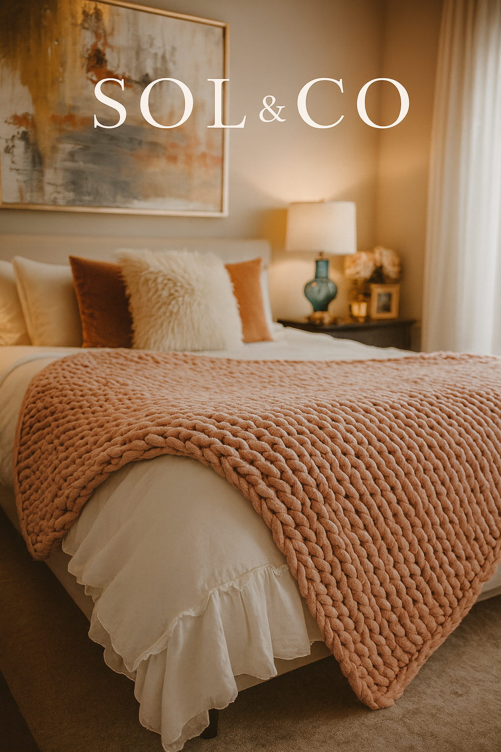 A cozy bedroom with a bed covered in a chunky pink blanket and white bedding. There are decorative pillows, including one fluffy white pillow and brown pillows. A nightstand holds a teal lamp, a framed picture, and flowers. A large abstract painting 