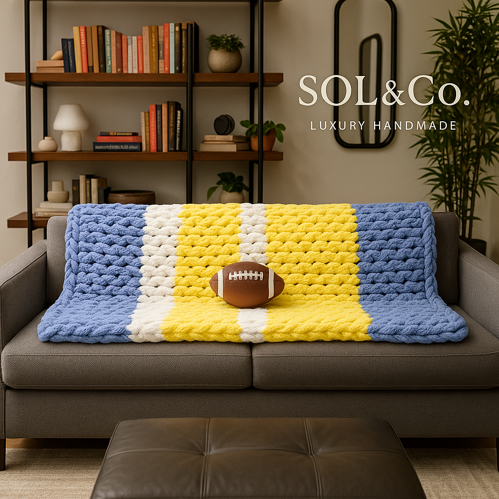 A gray sofa with a chunky knit blanket in blue, white, and yellow draped over it, with an American football placed on top of the blanket, situated in a cozy living room with a bookshelf, plants, and decorative items in the background.
