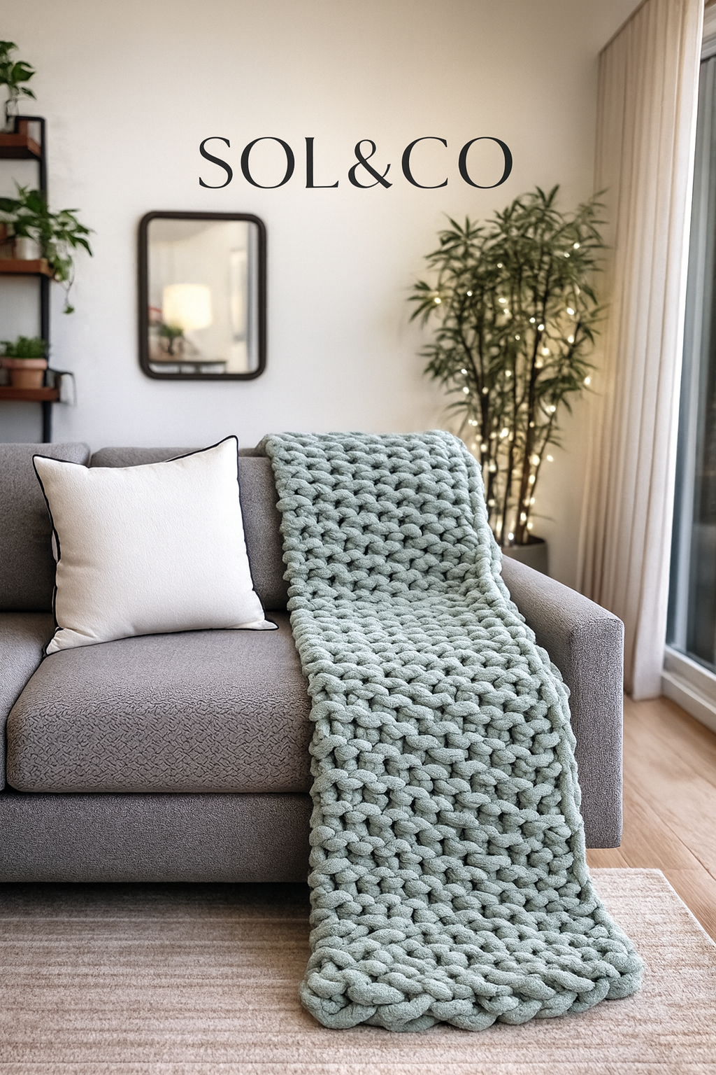 A living room with a grey sofa, a white pillow, a chunky knit blanket, a tall plant with string lights, a mirror, a wall shelf with plants, a rug, and a sign that reads 'SOL & CO' on the wall.