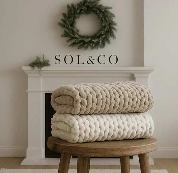Two chunky knitted blankets, one beige and one white, folded on a round wooden stool in front of a white mantel with a green wreath and the text 'SOL & CO' on the wall.