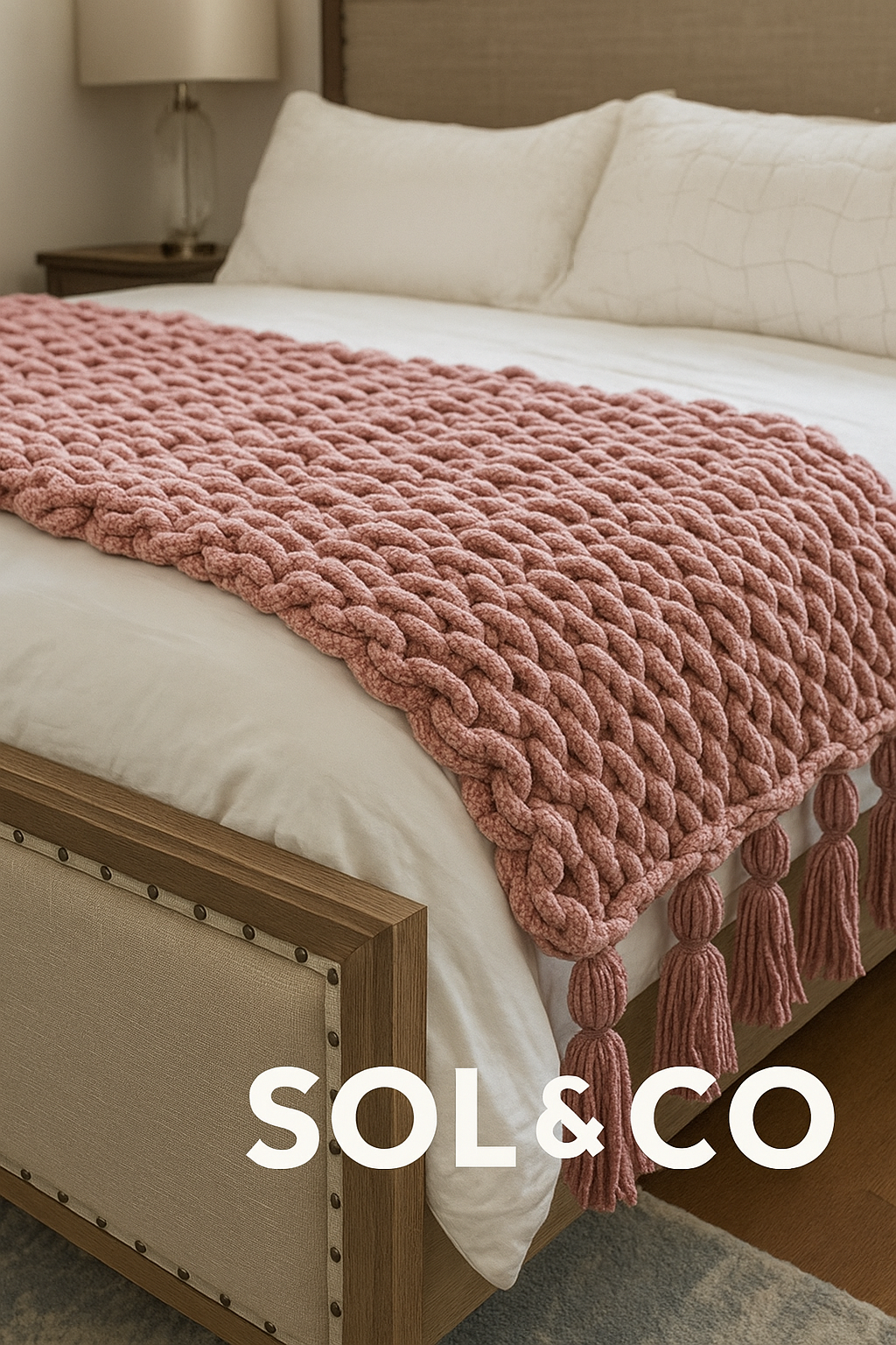 A bed with white bedding, a pink chunky knit blanket with tassels, beige pillows, a wooden headboard, and a nightstand with a lamp.