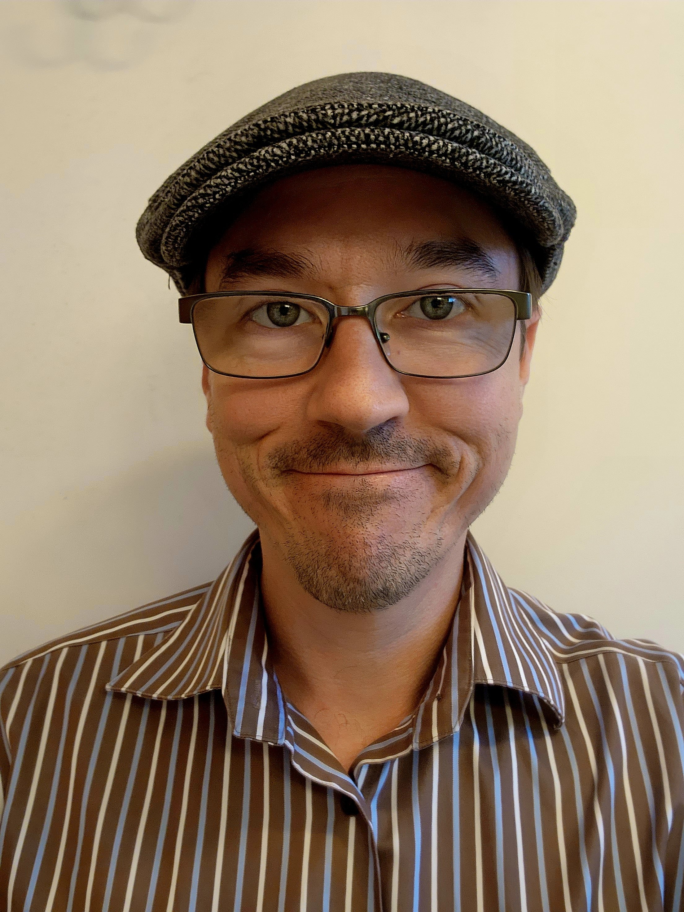 Close-up of Professor Brown wearing glasses, a gray cap, and a striped brown and white shirt, smiling at the camera against a plain background.