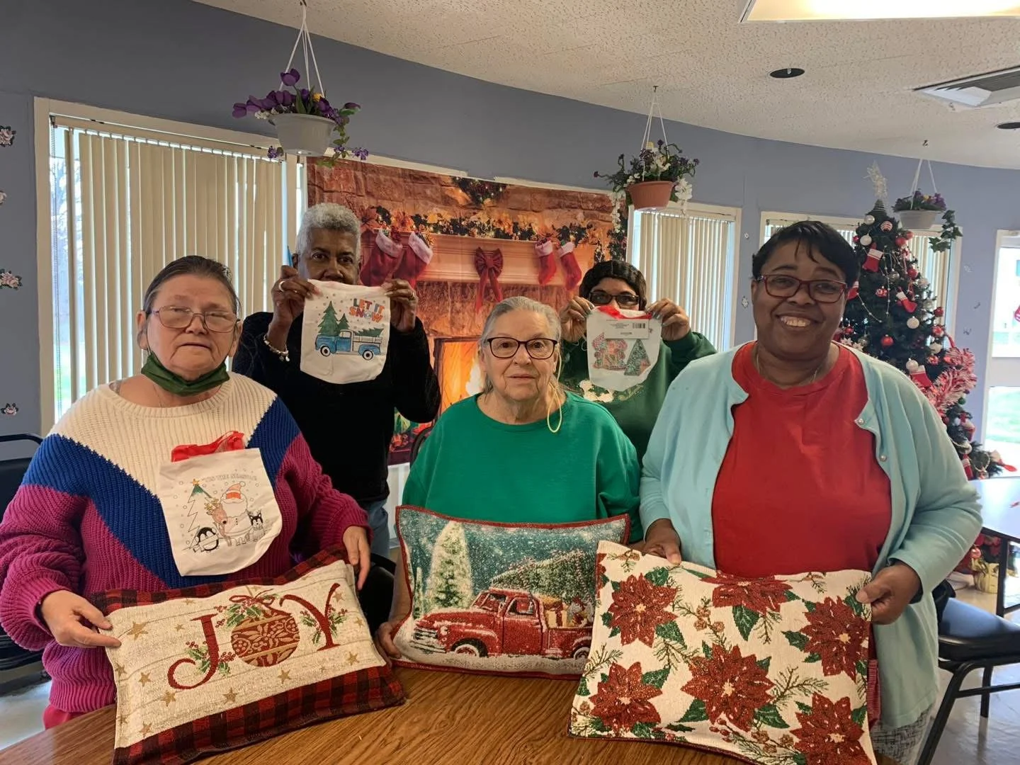 Five people, four women and one man, standing indoors during Christmas, holding decorative pillows and embroidered holiday towels. The background features a Christmas tree decorated with ornaments and a fireplace with stockings hanging, surrounded by festive decorations.
