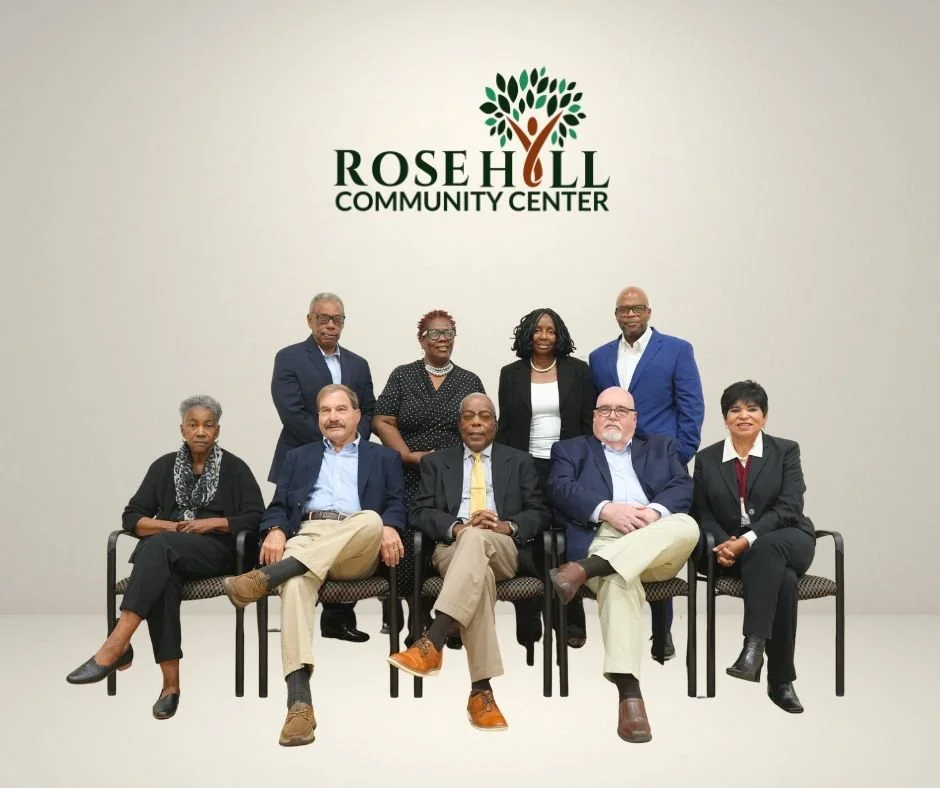 Group of nine diverse professionals posing in front of a wall with the Rose Hill Community Center logo, some seated and some standing.