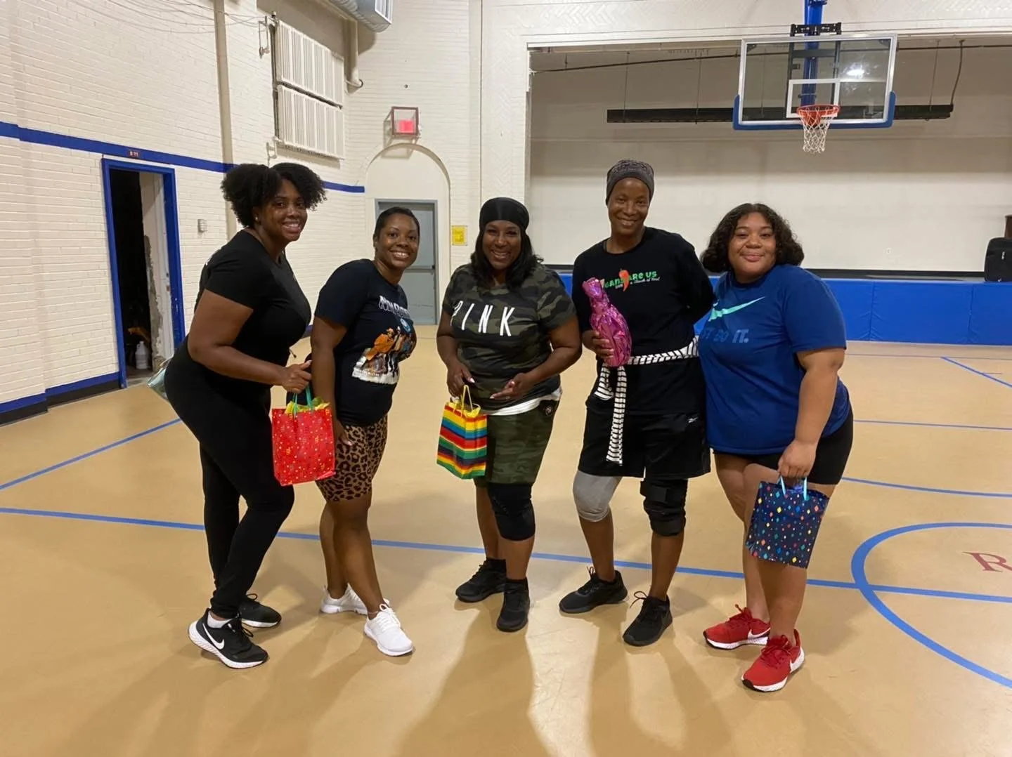 Five women standing in a gymnasium holding gift bags, smiling at the camera.