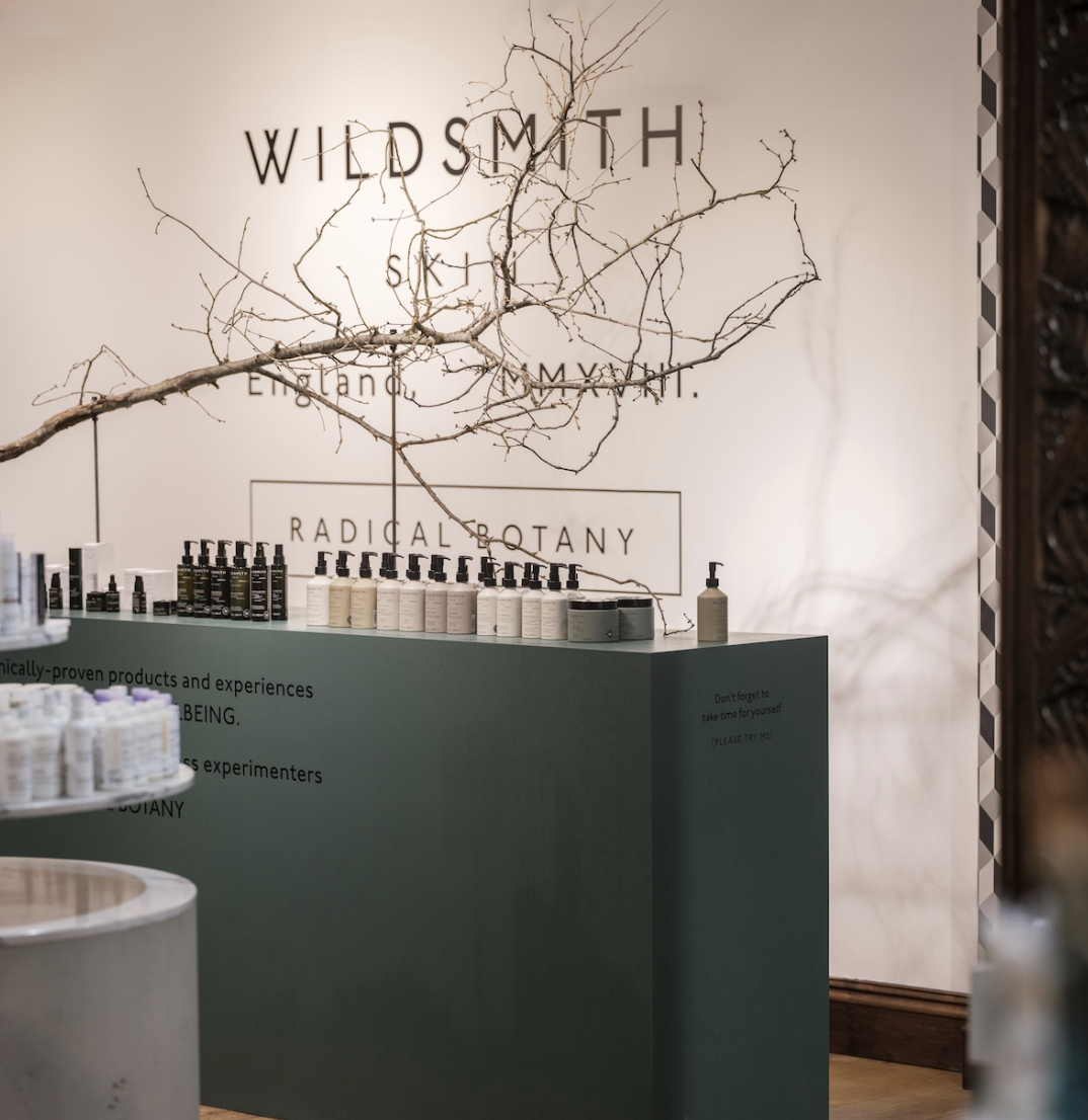 Display of skincare products on a green counter in a wellness spa or store with a sign reading 'Radical Botany' and a decorative tree branch on the wall.