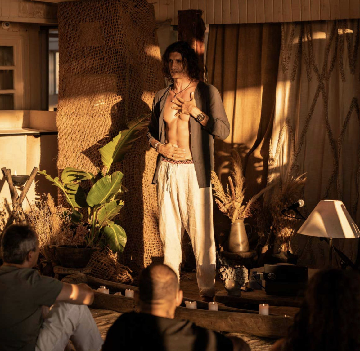 A man with long dark hair stands on a stage with a partially open shirt, revealing his chest and abdomen. He is surrounded by candles and plants, with warm lighting creating a dramatic atmosphere during a performance or presentation.