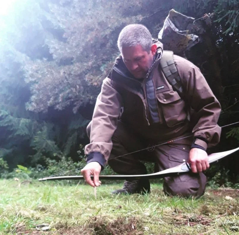 A man kneeling on the grass in a forest, examining the ground with a bow in his hand.