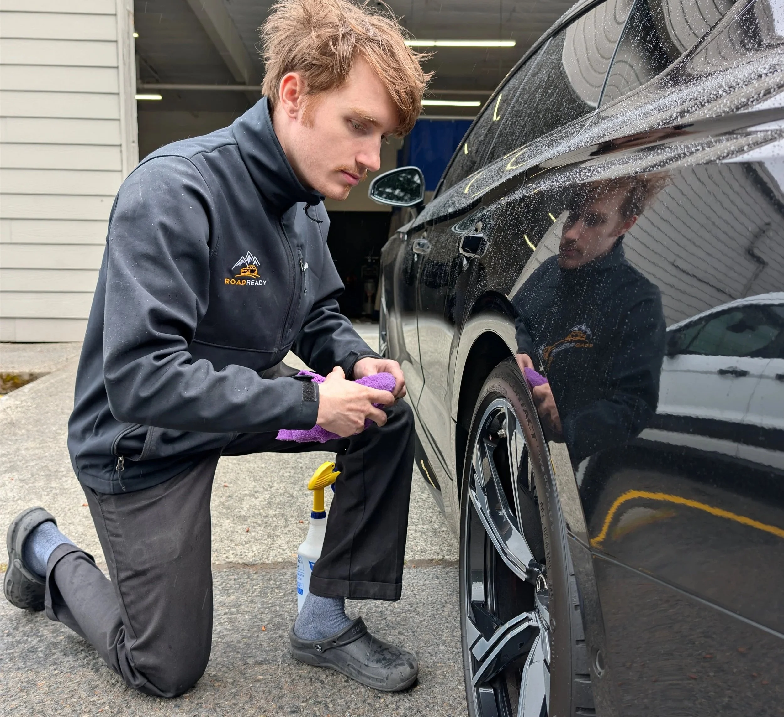 A young man kneeling outdoors in front of a black car, cleaning or detailing the car's tire with a purple cloth, wearing a gray jacket with a mountain logo and the words 'ROAD READY' on it.