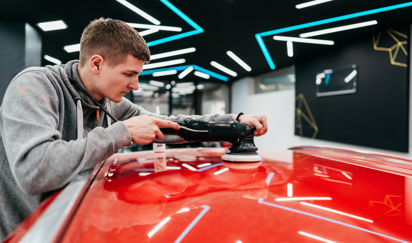 Young man polishing a red car with a power buffer inside a modern, well-lit auto detailing shop.