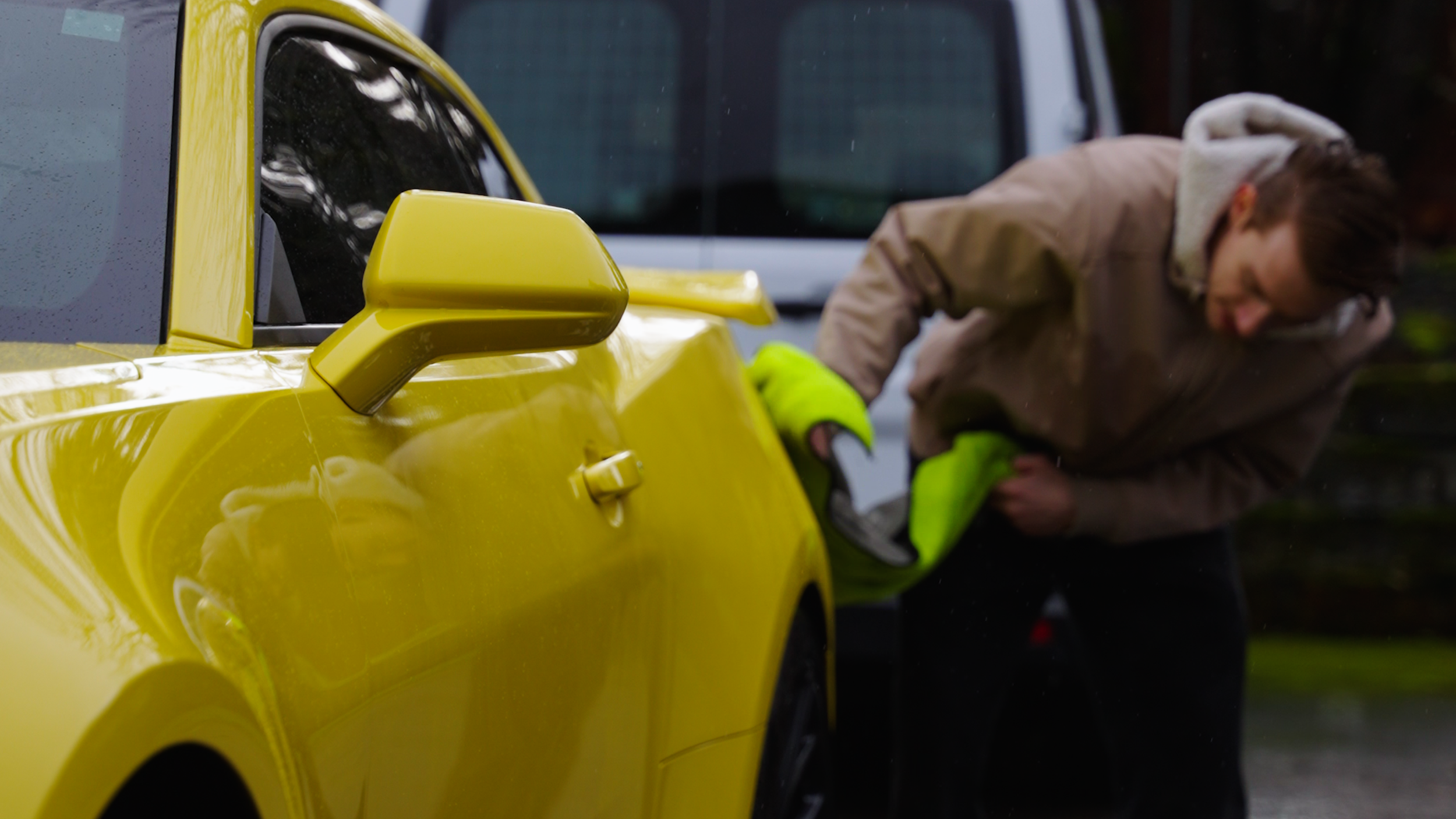 A person in a brown jacket and yellow gloves leaning into the driver's side of a bright yellow sports car, with a white van in the background.
