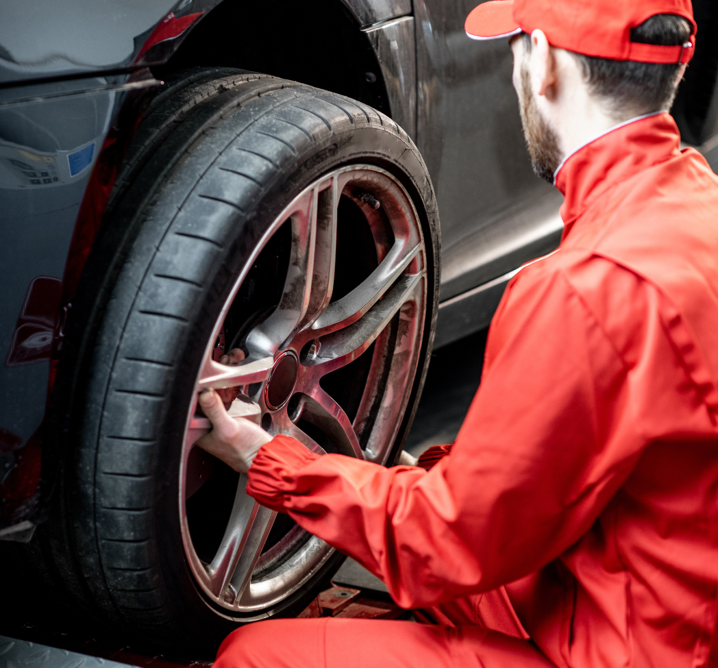 A mechanic in red coveralls and cap checking a car's wheel and tire in a garage.
