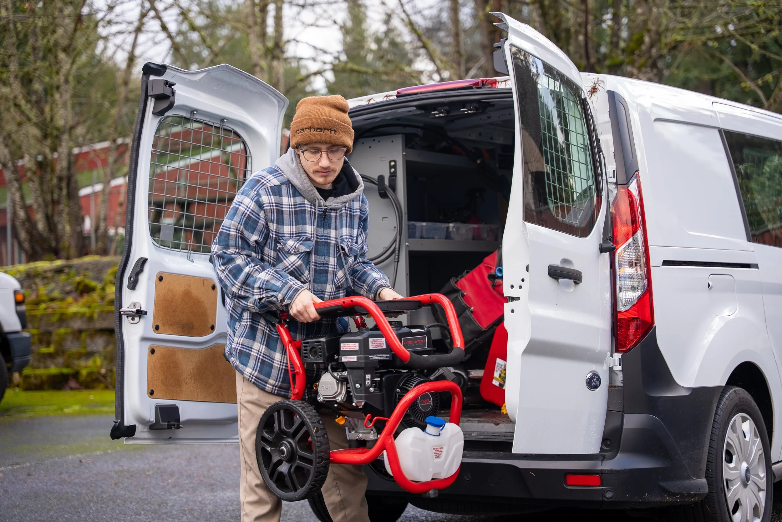 A man in a plaid shirt, beige pants, glasses, and a brown beanie, working with a generator outside a white van. The van's back doors are open, revealing tools and equipment inside. The man is holding the generator and standing on a paved road with tr
