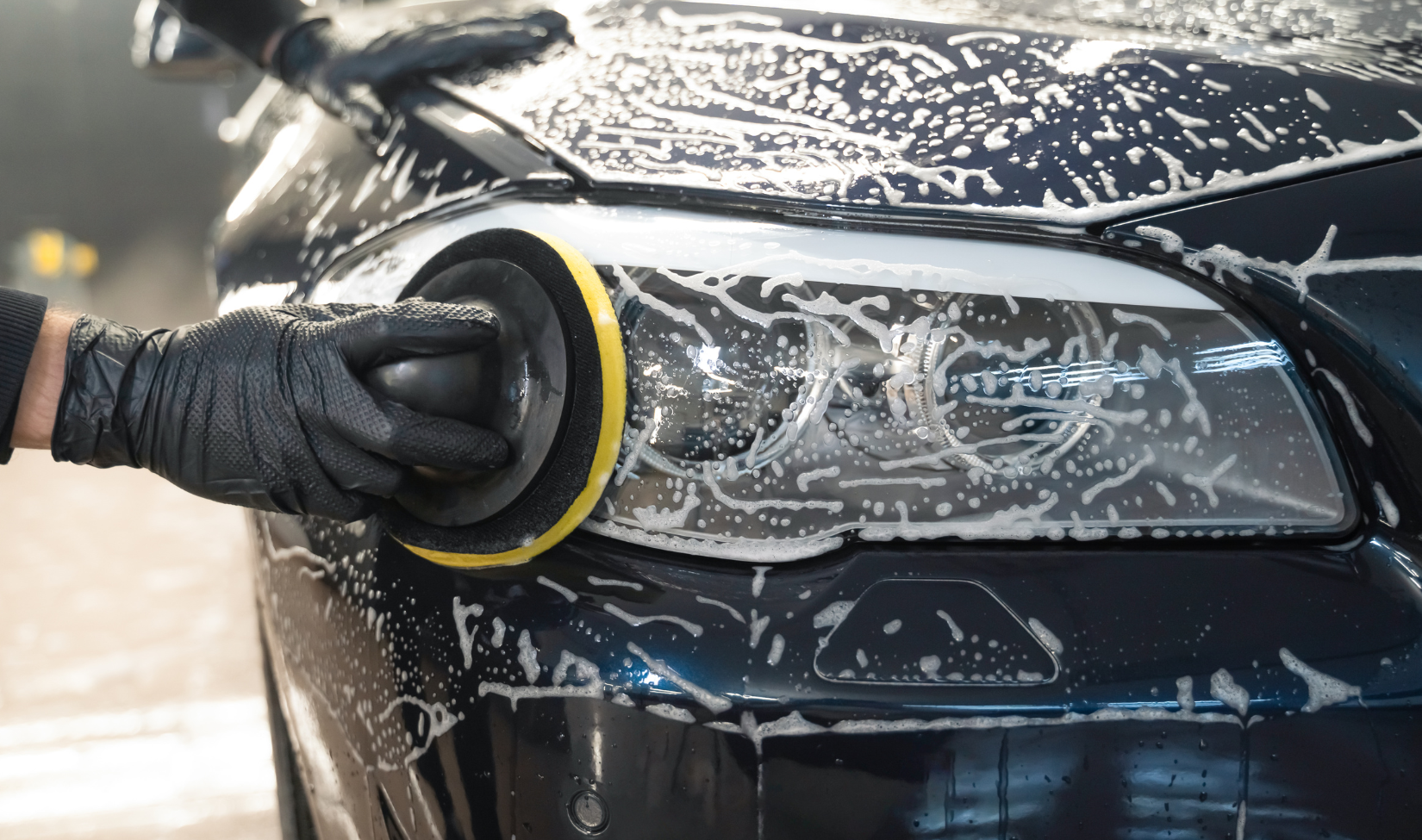 Person wearing black gloves using a machine polisher to clean the headlight of a black car, with soap and foam.