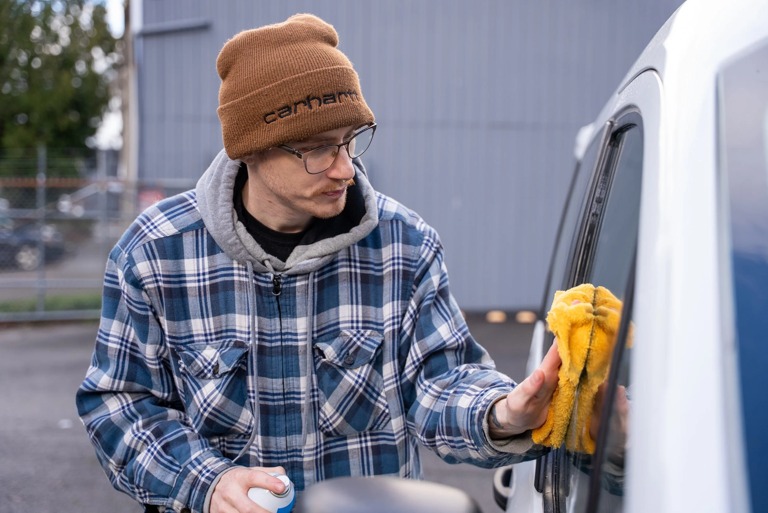 A man wearing a brown beanie, glasses, a gray hoodie, and a blue plaid shirt is cleaning the side of a white vehicle with a yellow cloth, holding a spray bottle in his other hand.