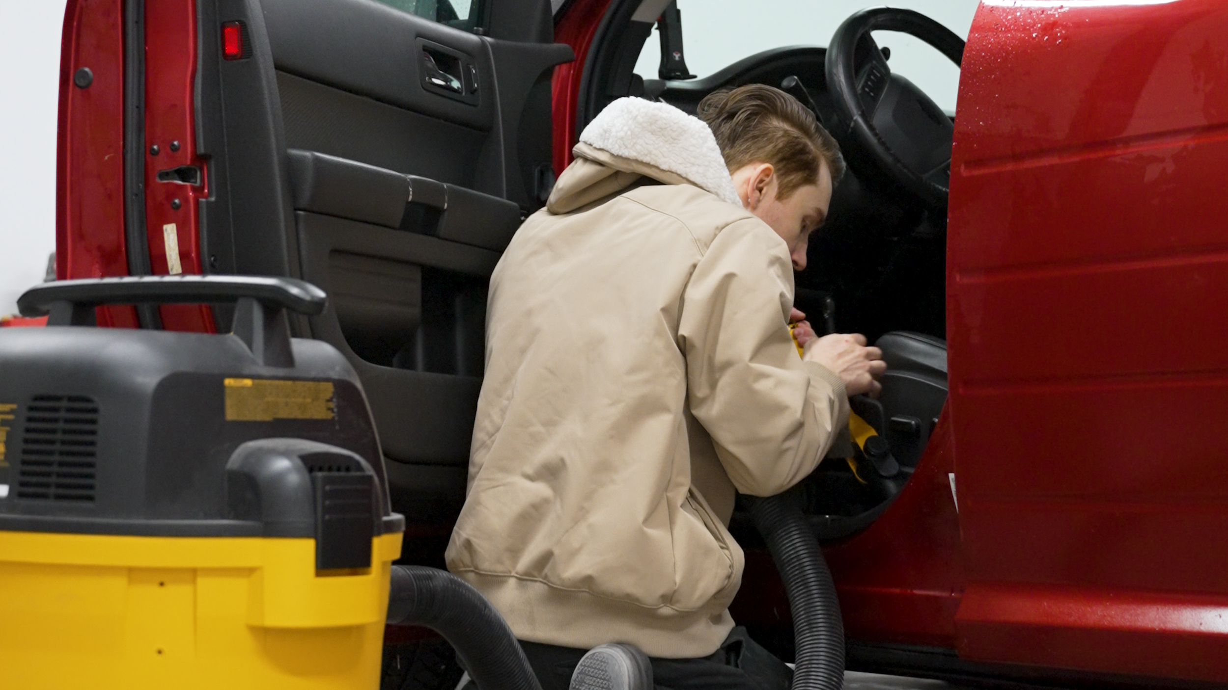 A man wearing a beige jacket and a white fleece hoodie working on the interior of a red vehicle, possibly a fire truck, with a yellow industrial vacuum and other equipment nearby.