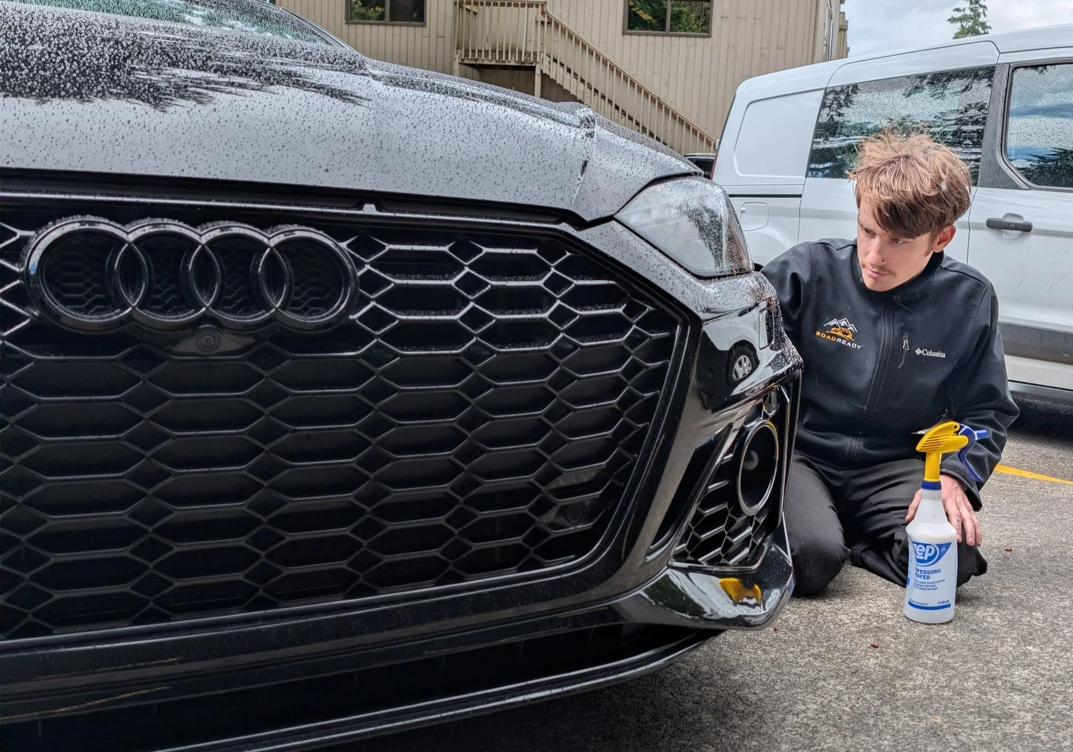A man is cleaning the front of a black Audi car, using a spray bottle and a yellow cloth, with water droplets visible on the car's surface and surroundings, and a white van in the background.