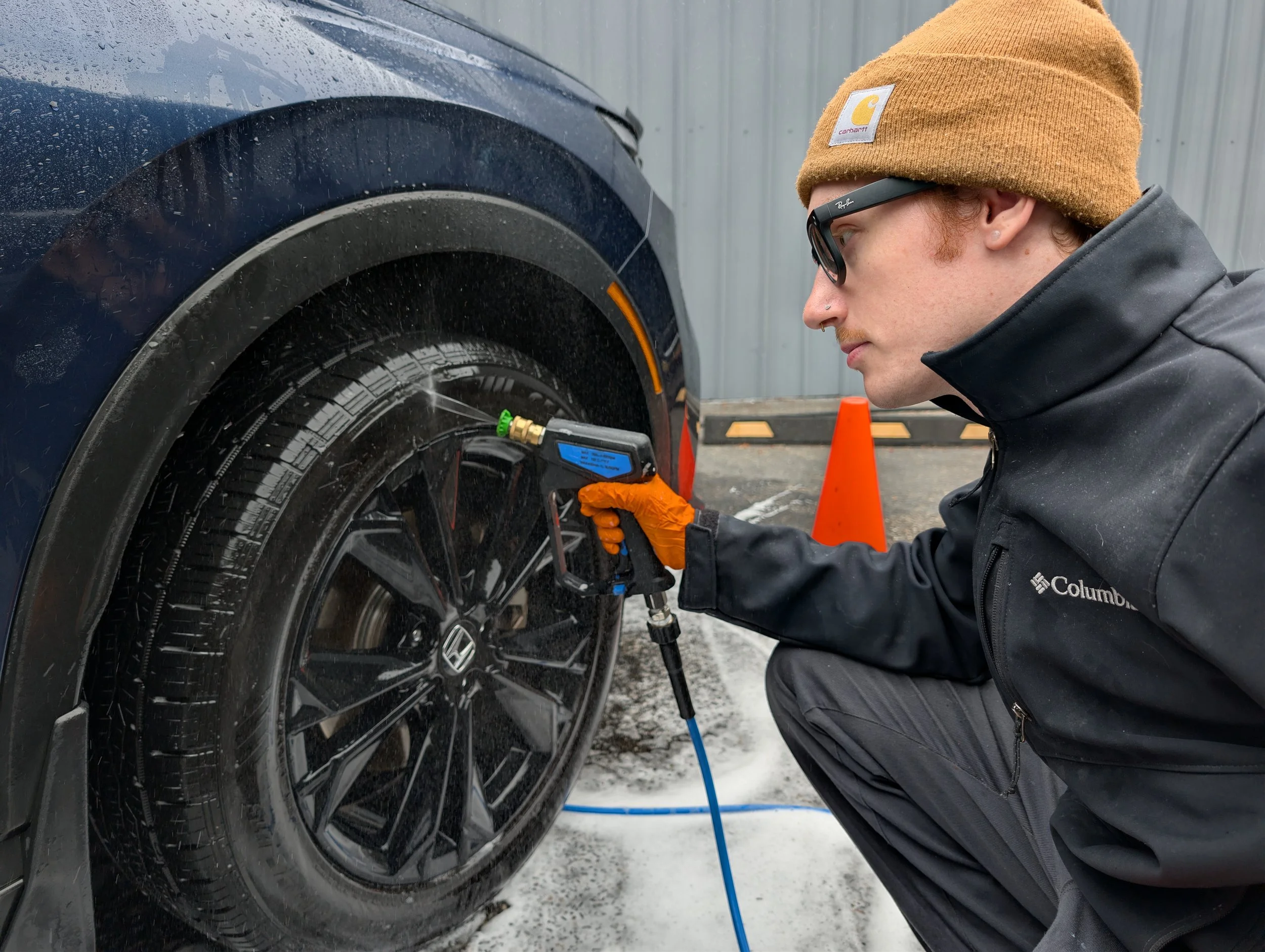A person in a brown beanie, glasses, and a black jacket inflates a car tire with a portable air compressor, with orange safety cones behind him and snow on the ground.