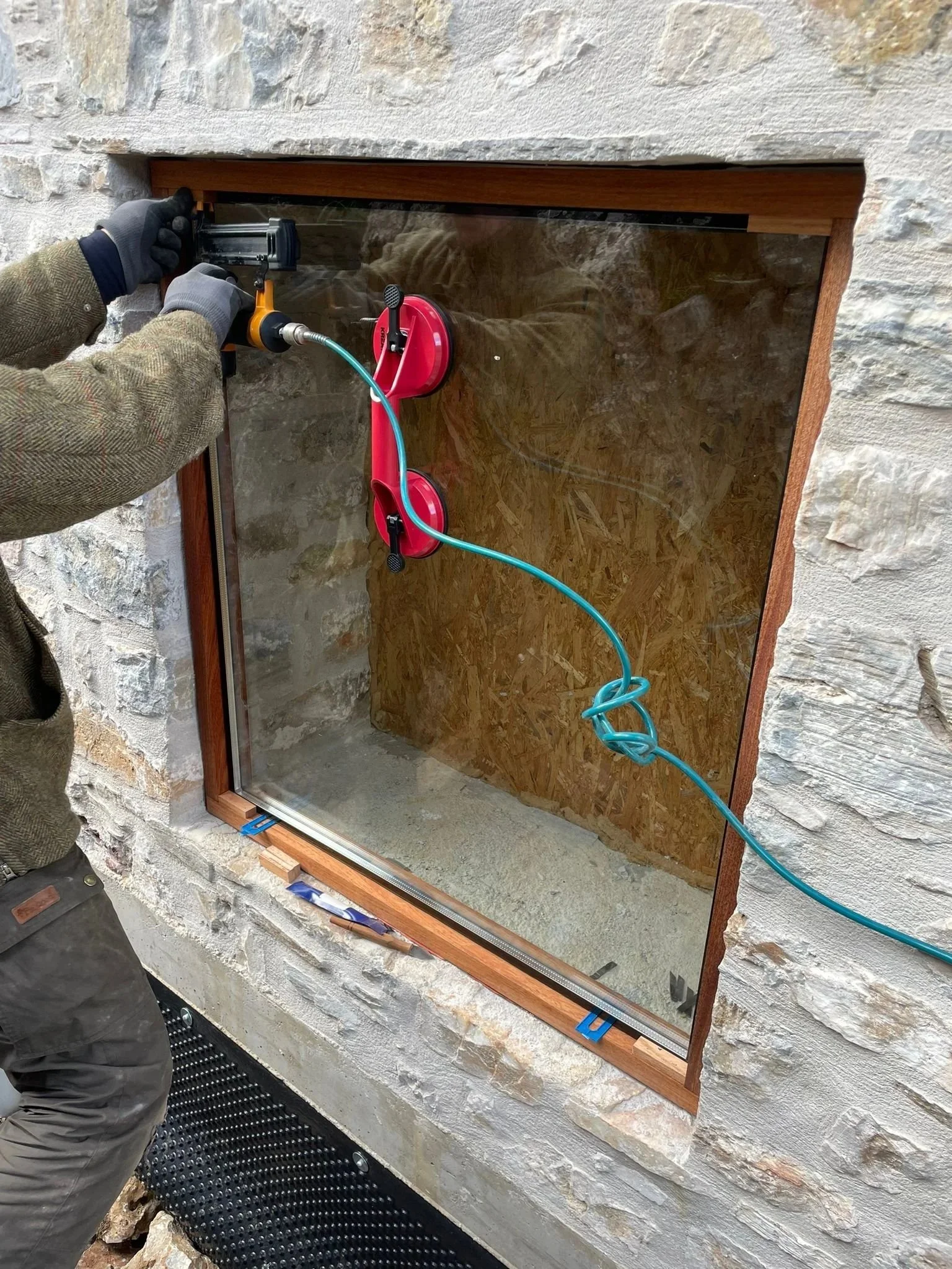 Person installing a glass window in a stone wall using a caulking gun with a blue cord.