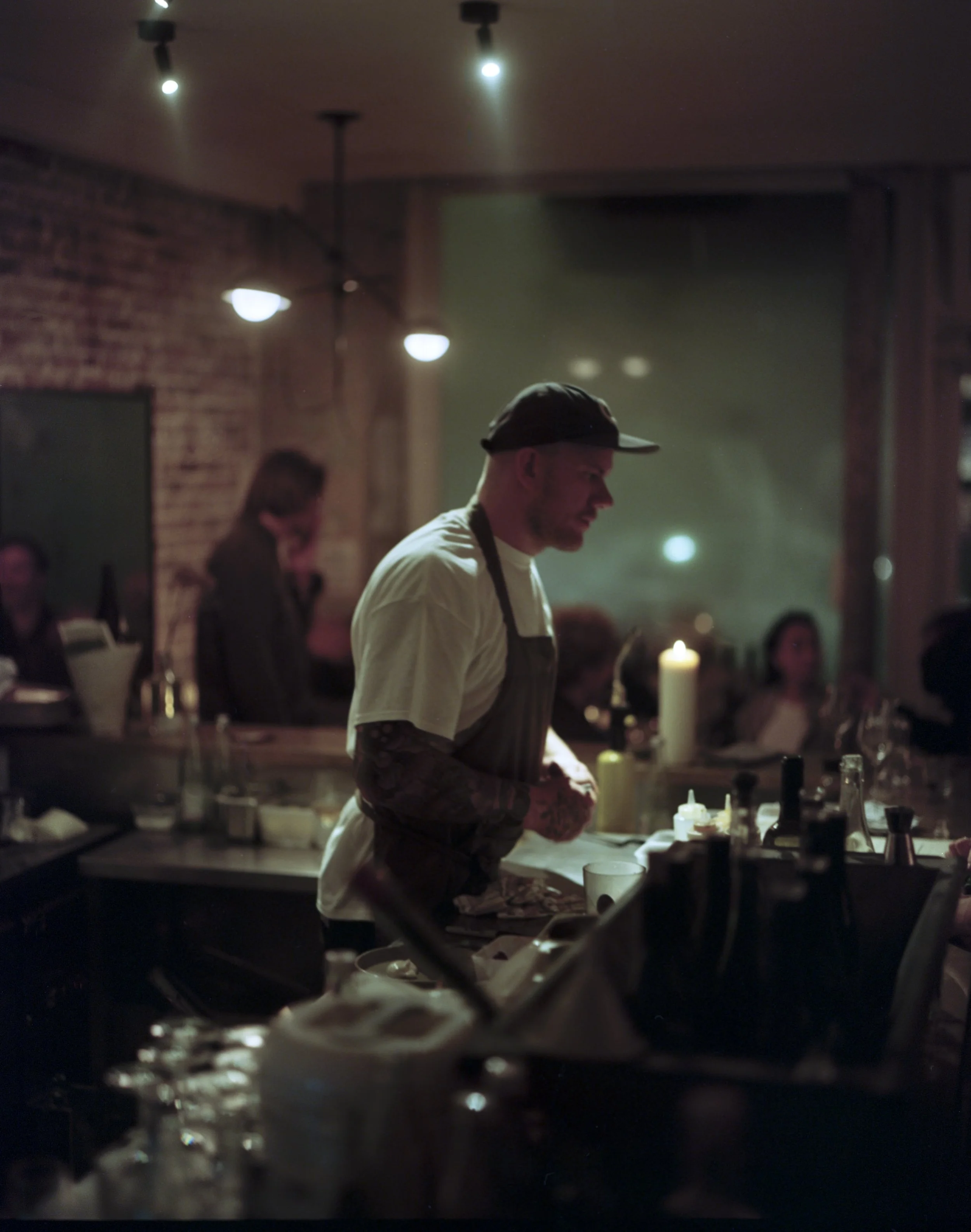 A bartender with tattooed arms, wearing a cap and apron, preparing drinks in a dimly lit bar with other patrons in the background.