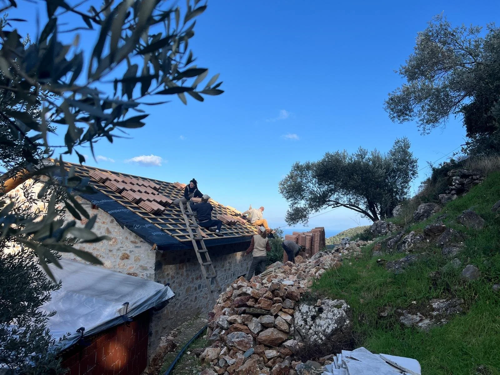 Construction workers installing roof tiles on a stone building in a hilly, green landscape under a blue sky.