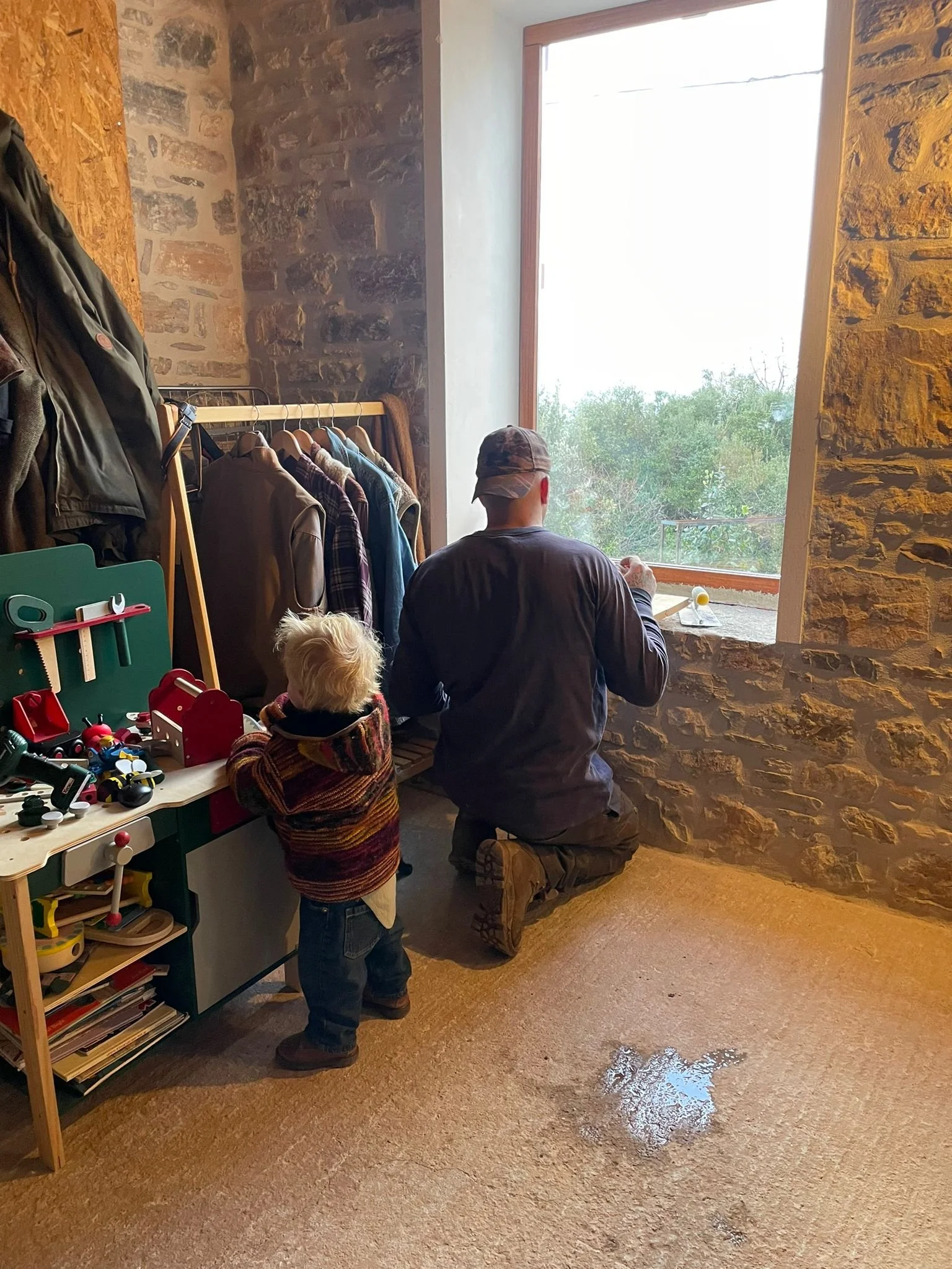 A man and a young boy looking out of a large window with stone walls inside a rustic room. The boy is standing near a toy shelf with toys, and the man is kneeling by the windowsill.