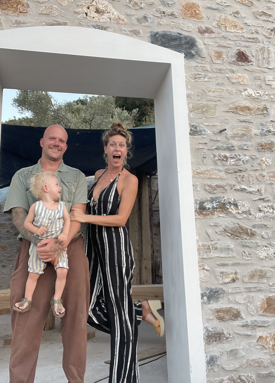 A man with tattoos on his arm holding a young girl in front of a woman, all smiling and making playful expressions, standing outside between stone walls and a large white doorway.