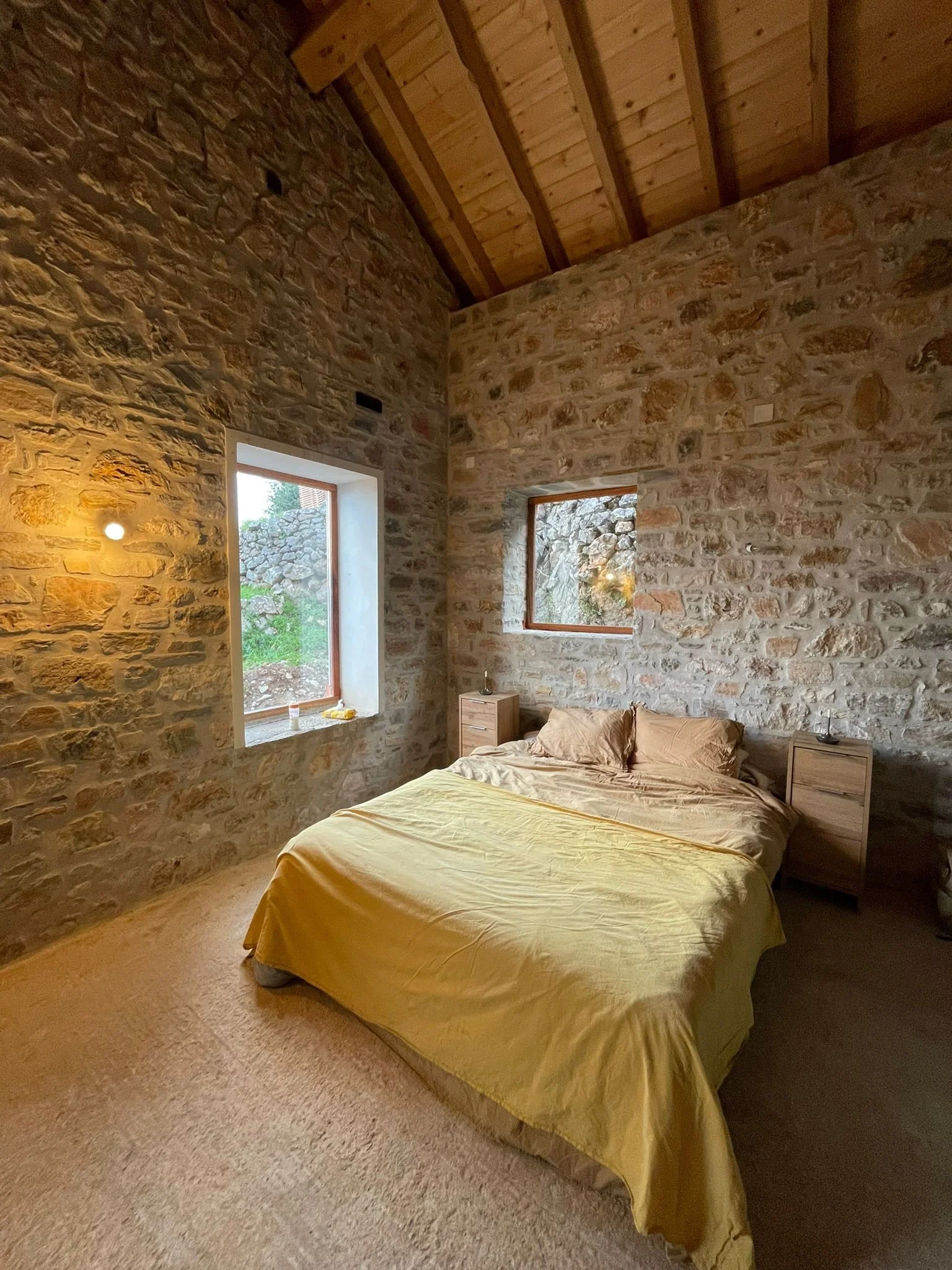 A cozy bedroom with stone walls, two small windows, a bed with beige bedding and matching pillows, and two wooden nightstands.