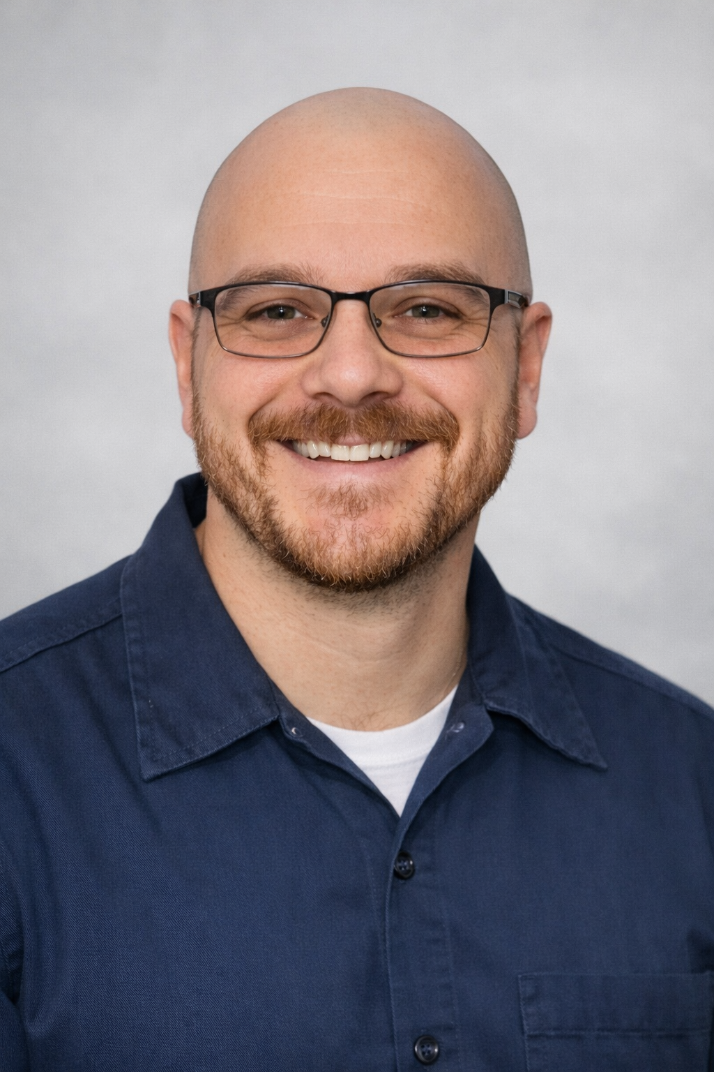 A smiling man with glasses, a beard, and a mustache, wearing a dark blue collared shirt against a plain light gray background.