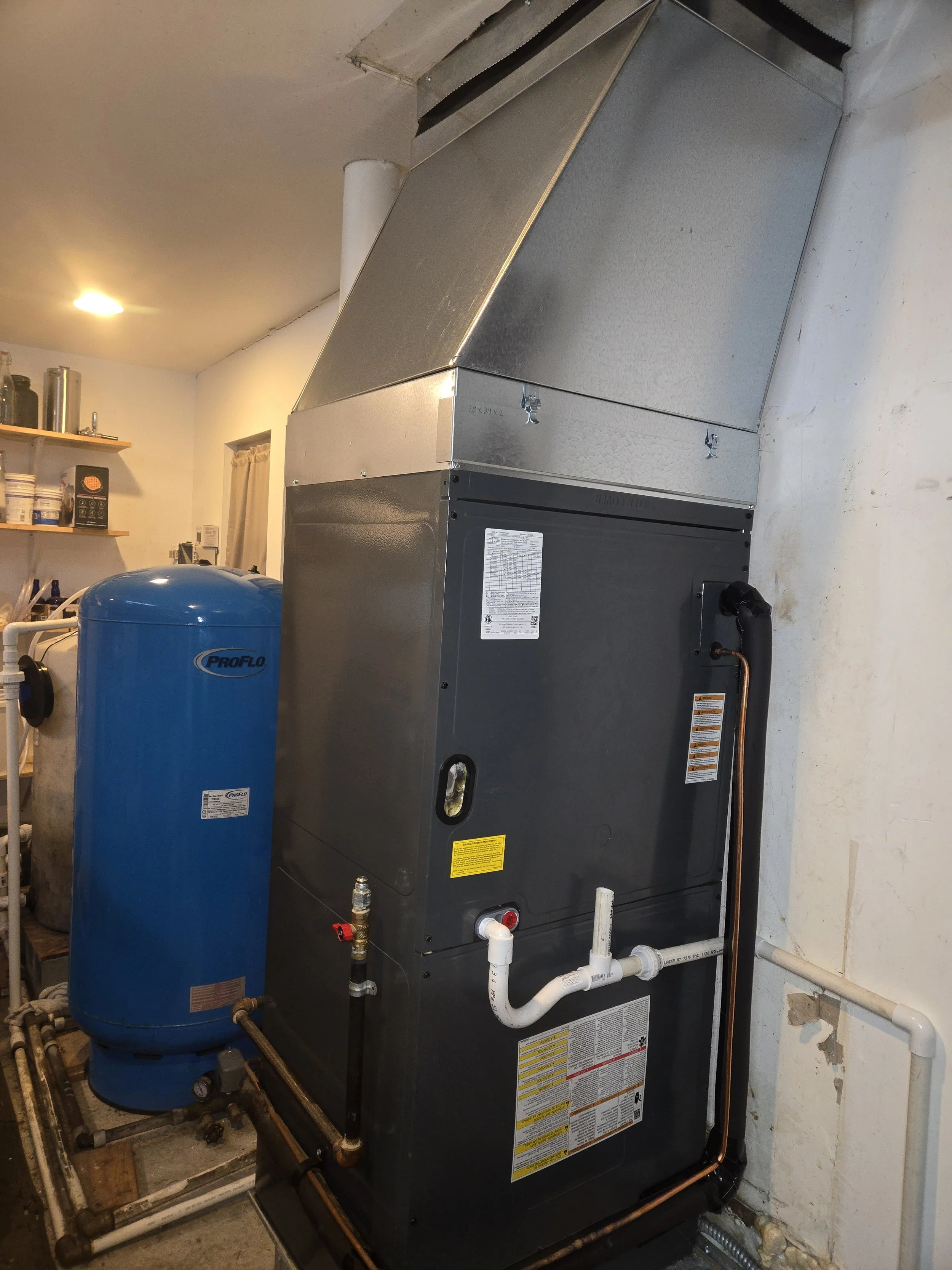 A large gray and black HVAC furnace with white pipes and copper tubing in a utility room. A blue water pressure tank labeled "Pro Flo" is nearby, with shelves containing various supplies in the background.