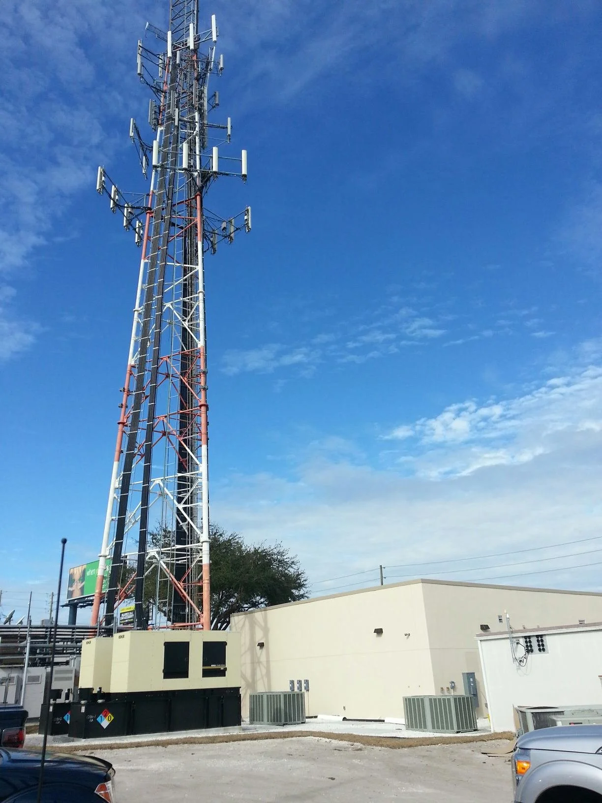 A tall telecommunication tower with antennas stands against a blue sky with scattered clouds, adjacent to a beige building and parked cars.