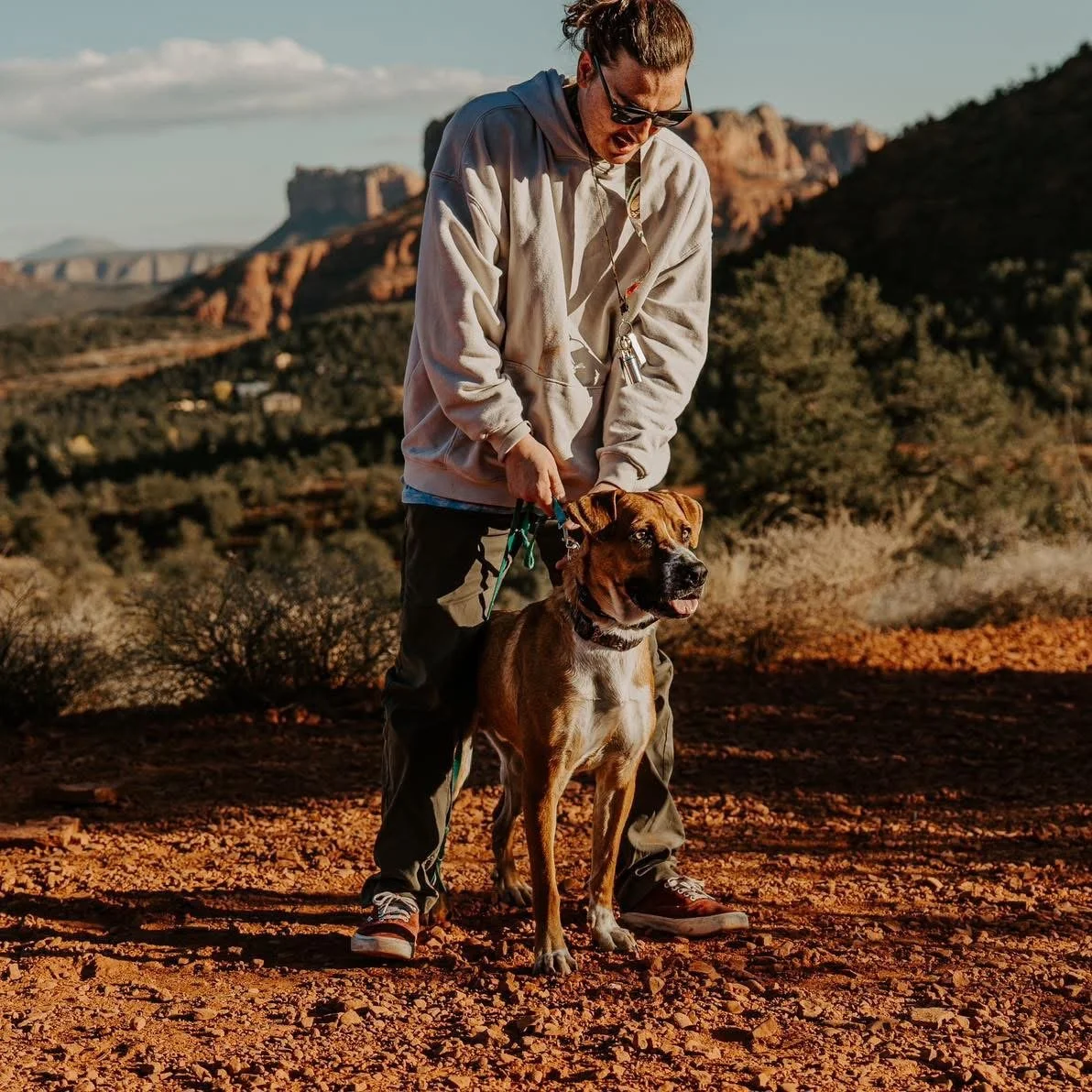 A person in sunglasses and a hoodie holding a leash on a brown and white dog in a rocky desert landscape.