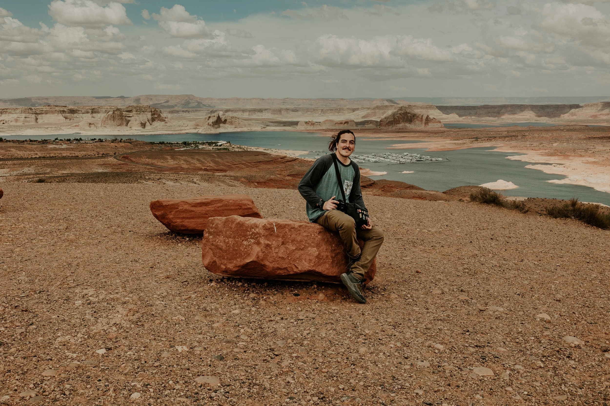 A person with a camera sitting on large red rocks, overlooking a river with a marina, surrounded by desert landscape and distant canyon formations under a cloudy sky. photographer Pablo Cruz sitting on rocks in Glen Canyon National Park