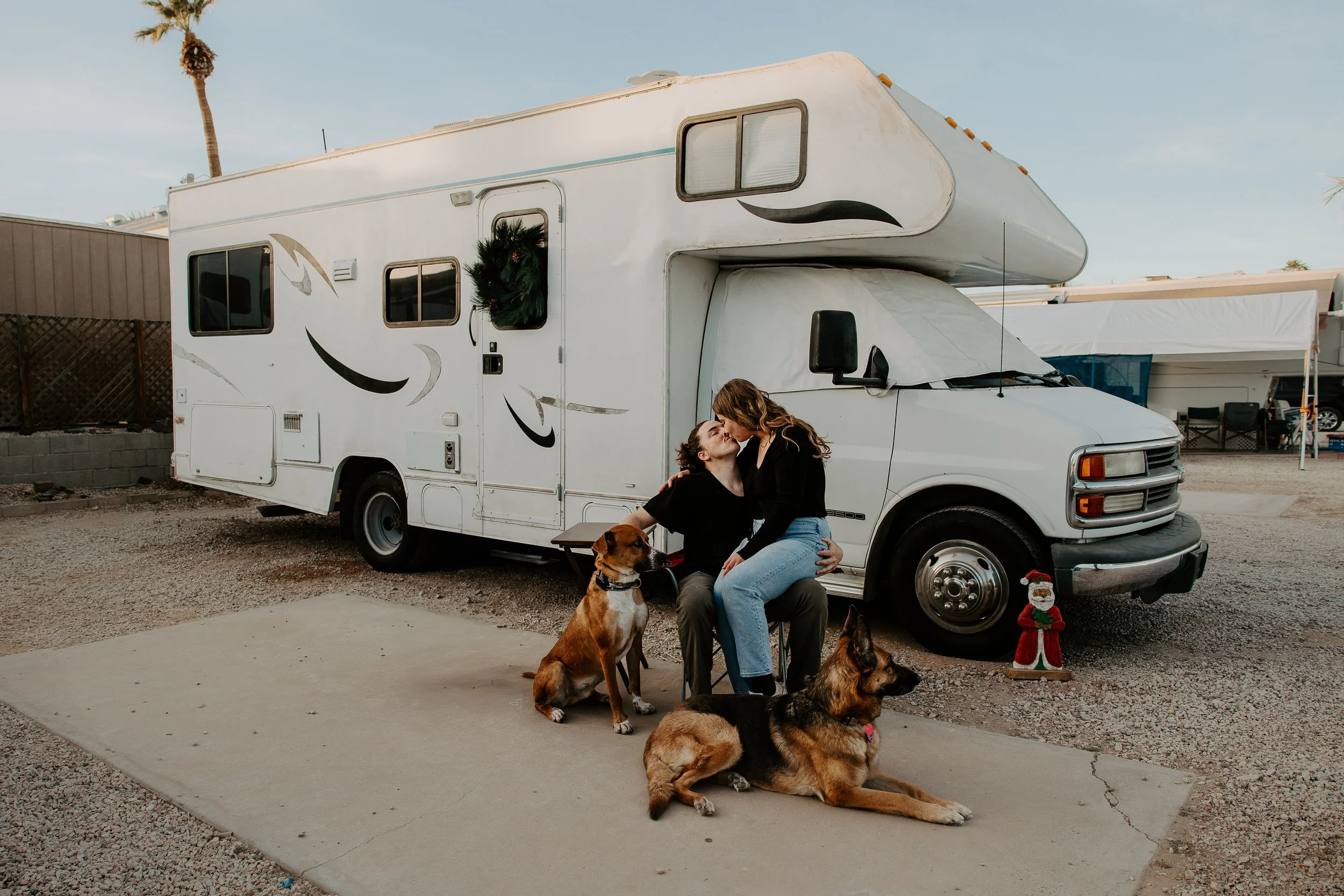 A couple and two dogs sit in front of a white camper van, sharing a kiss. The woman is seated, and the man is sitting on a chair. The scene is decorated with a small Christmas figure and a wreath on the camper.