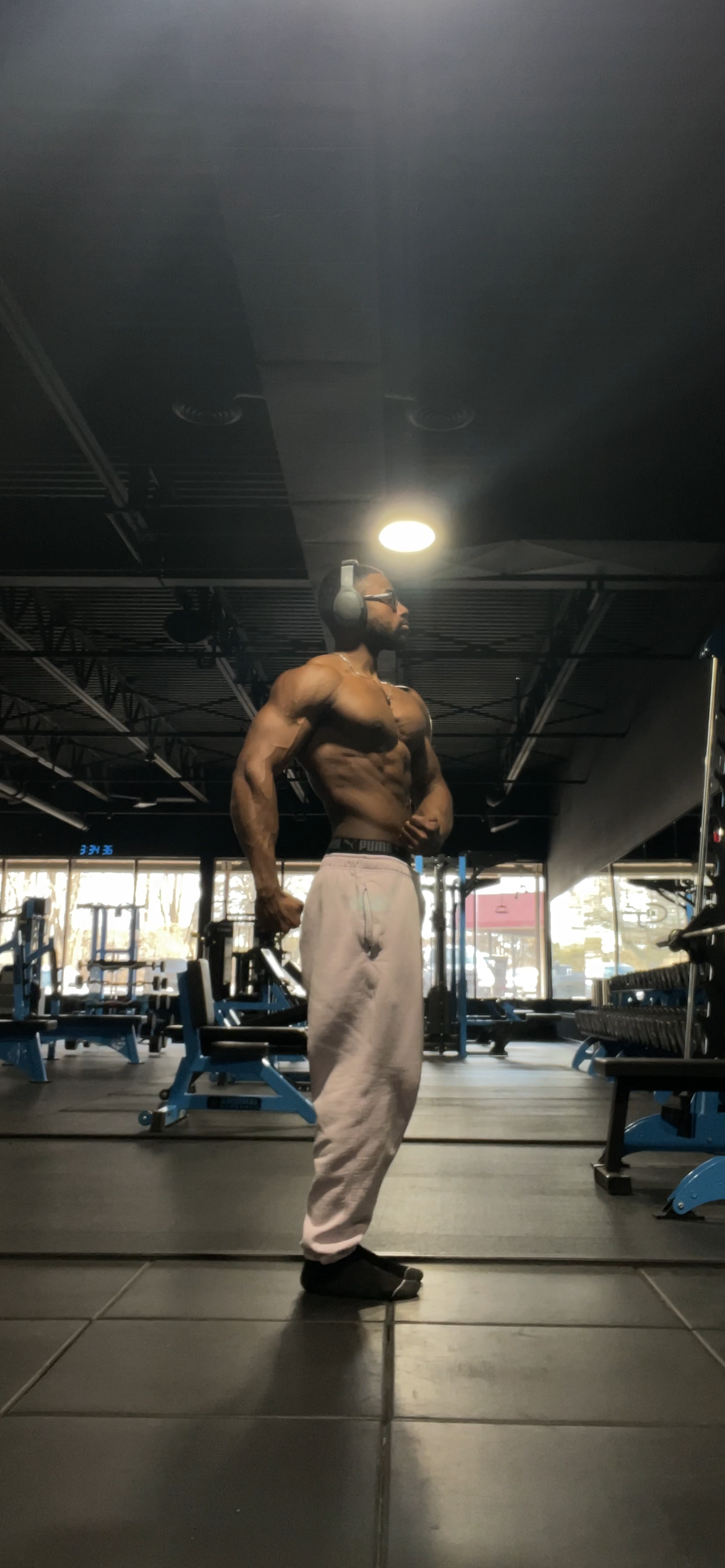 A muscular man wearing headphones and glasses, shirtless, in a gym, standing and posing with his left arm flexed, wearing gray sweatpants and black socks.