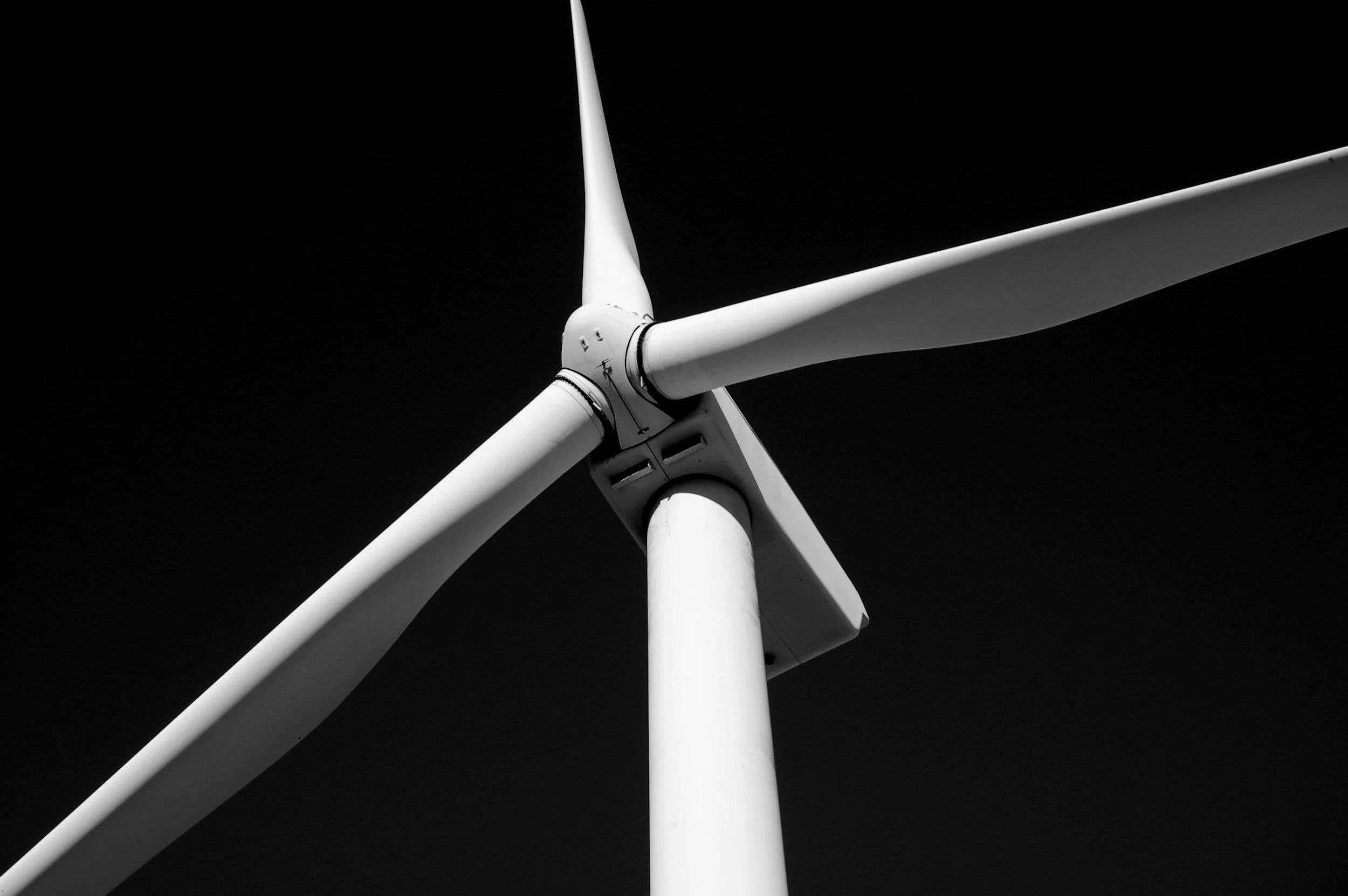A black and white photograph of a wind turbine taken from below, showing the blades and hub against a dark sky.