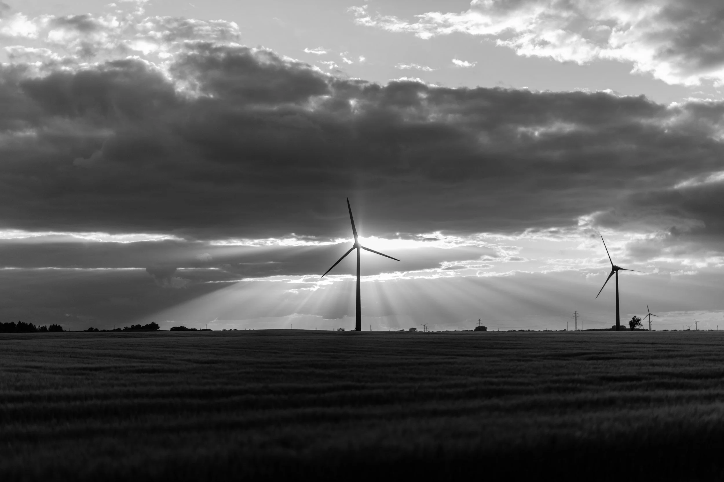 Black and white photograph of wind turbines standing in a field under a sky filled with dark clouds, with the sun setting behind the clouds and rays of sunlight shining through.