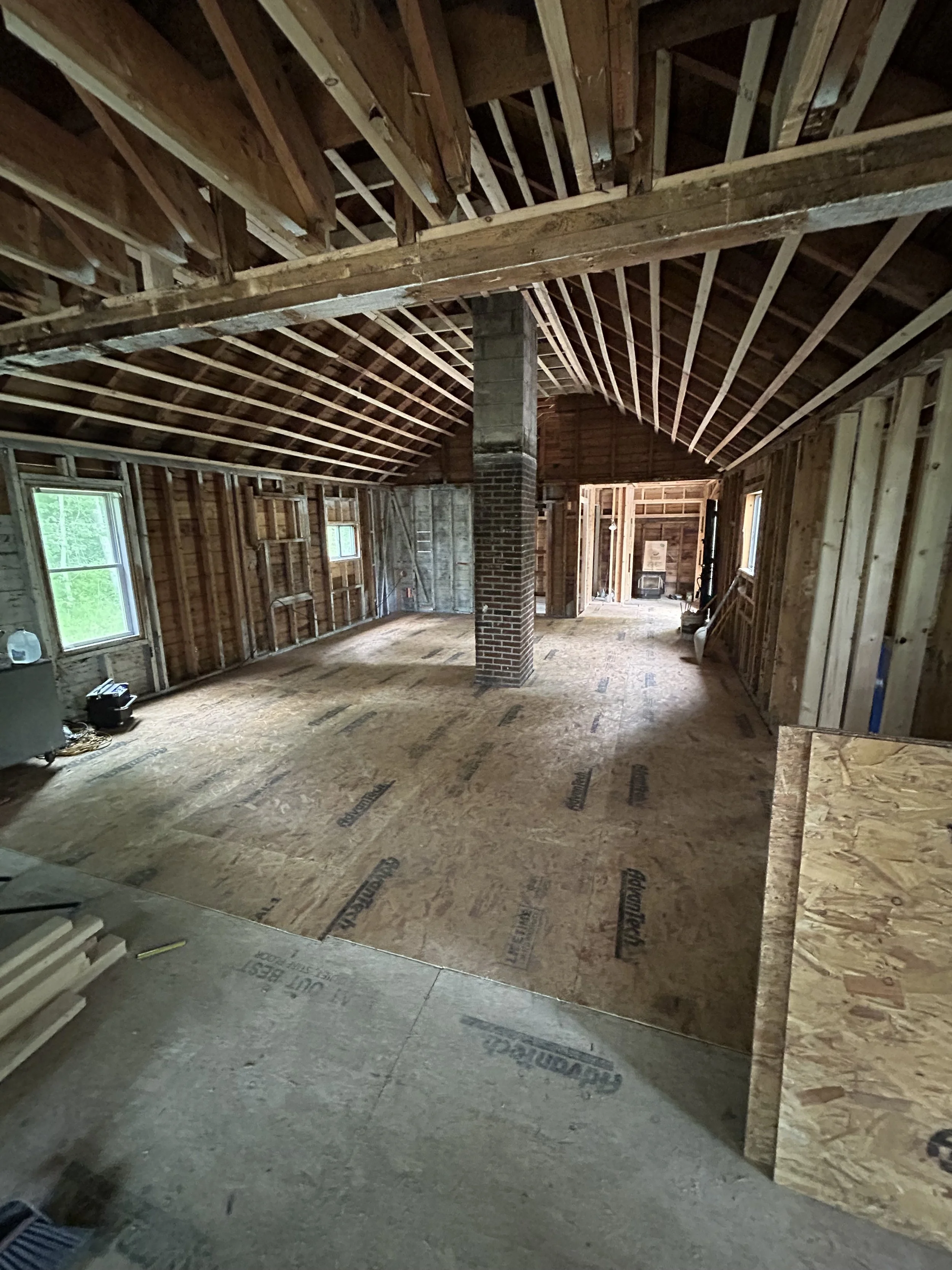Interior of a house under construction, showing exposed wooden framing, a brick chimney, and plywood subflooring.