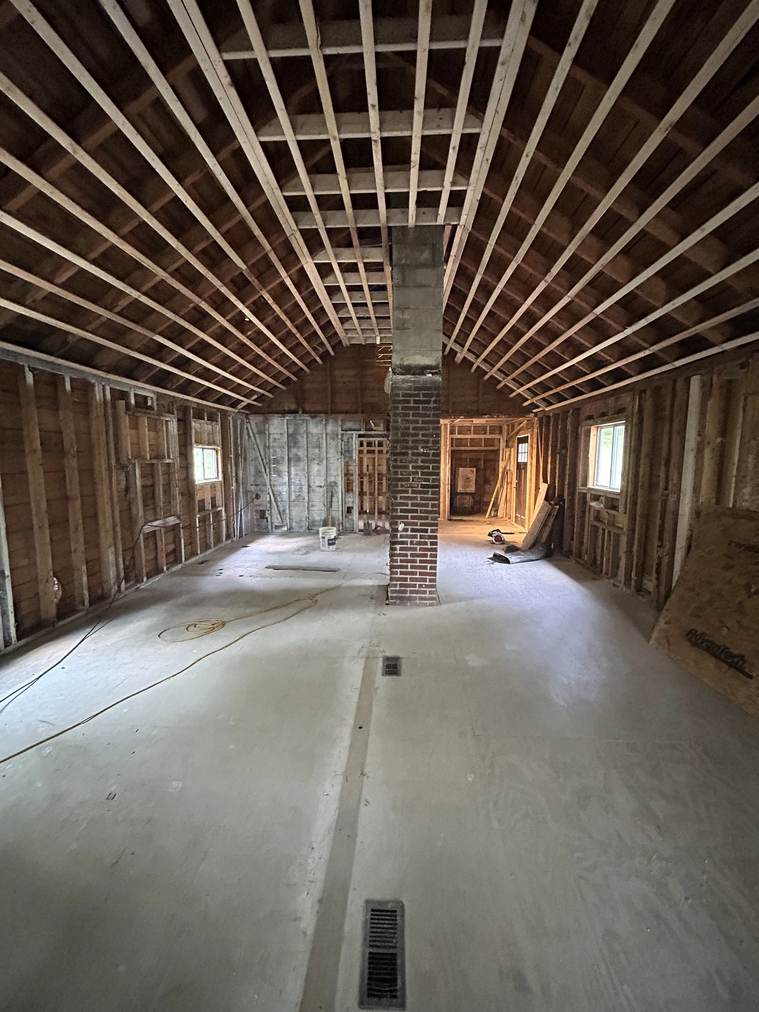 Interior of a house under construction with exposed wooden framing, a brick chimney in the center, and two small windows on each side.