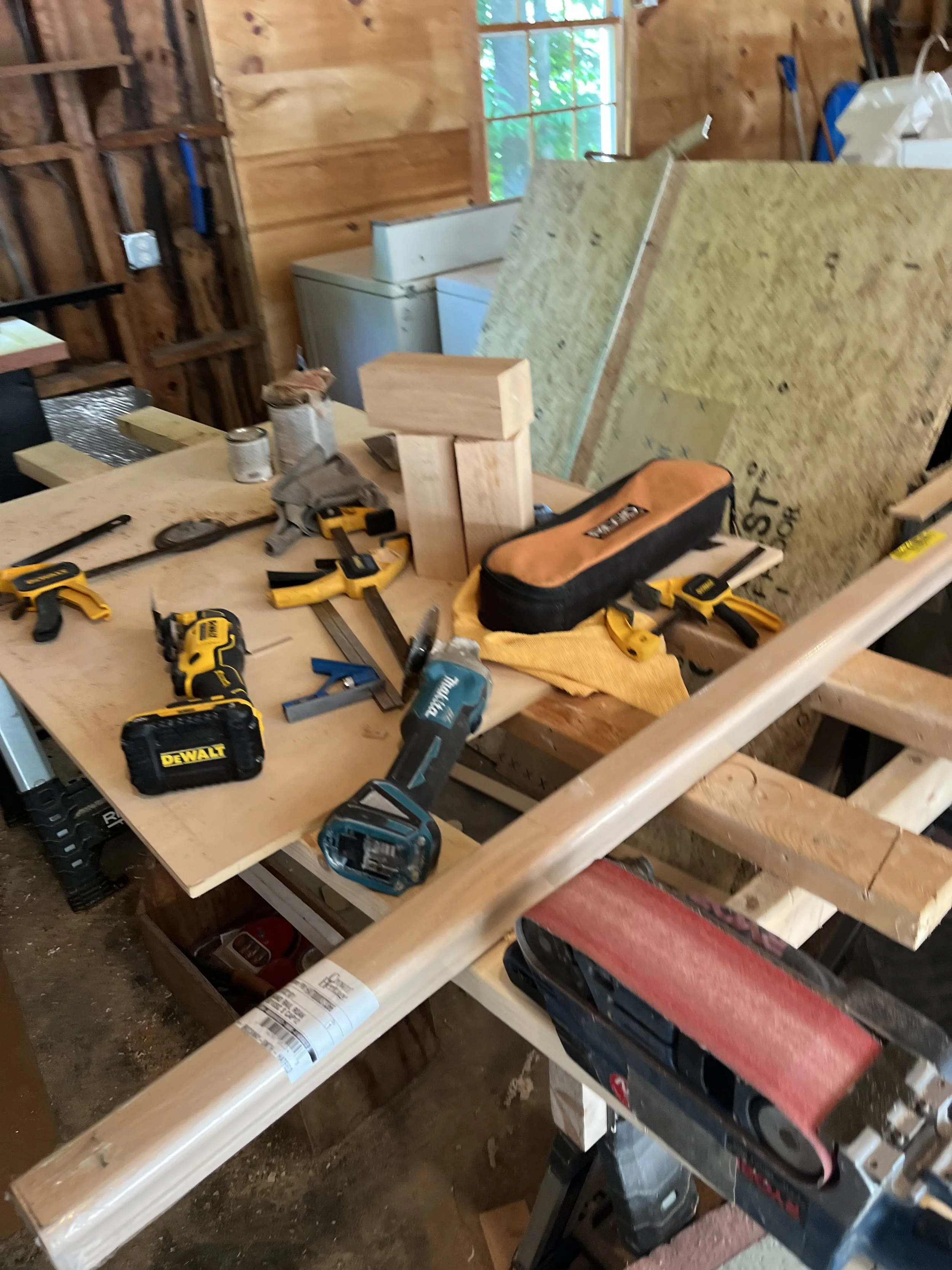 A woodworking workspace with various power tools, wooden pieces, clamps, and sheets of insulation or plywood on a workbench.
