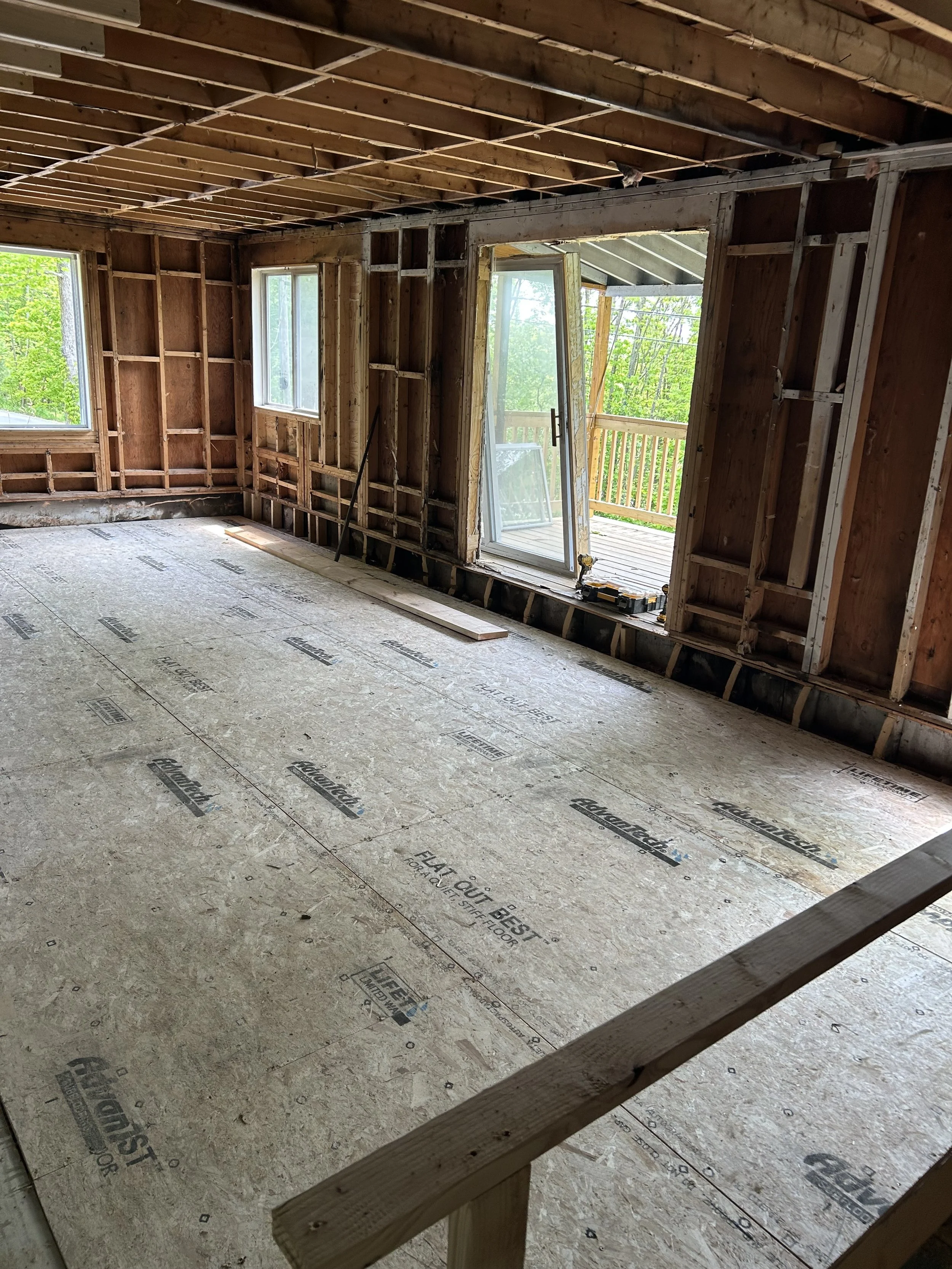 Interior of a house under construction with open walls, windows, and a door leading to an outdoor deck, showing exposed framing and new flooring.