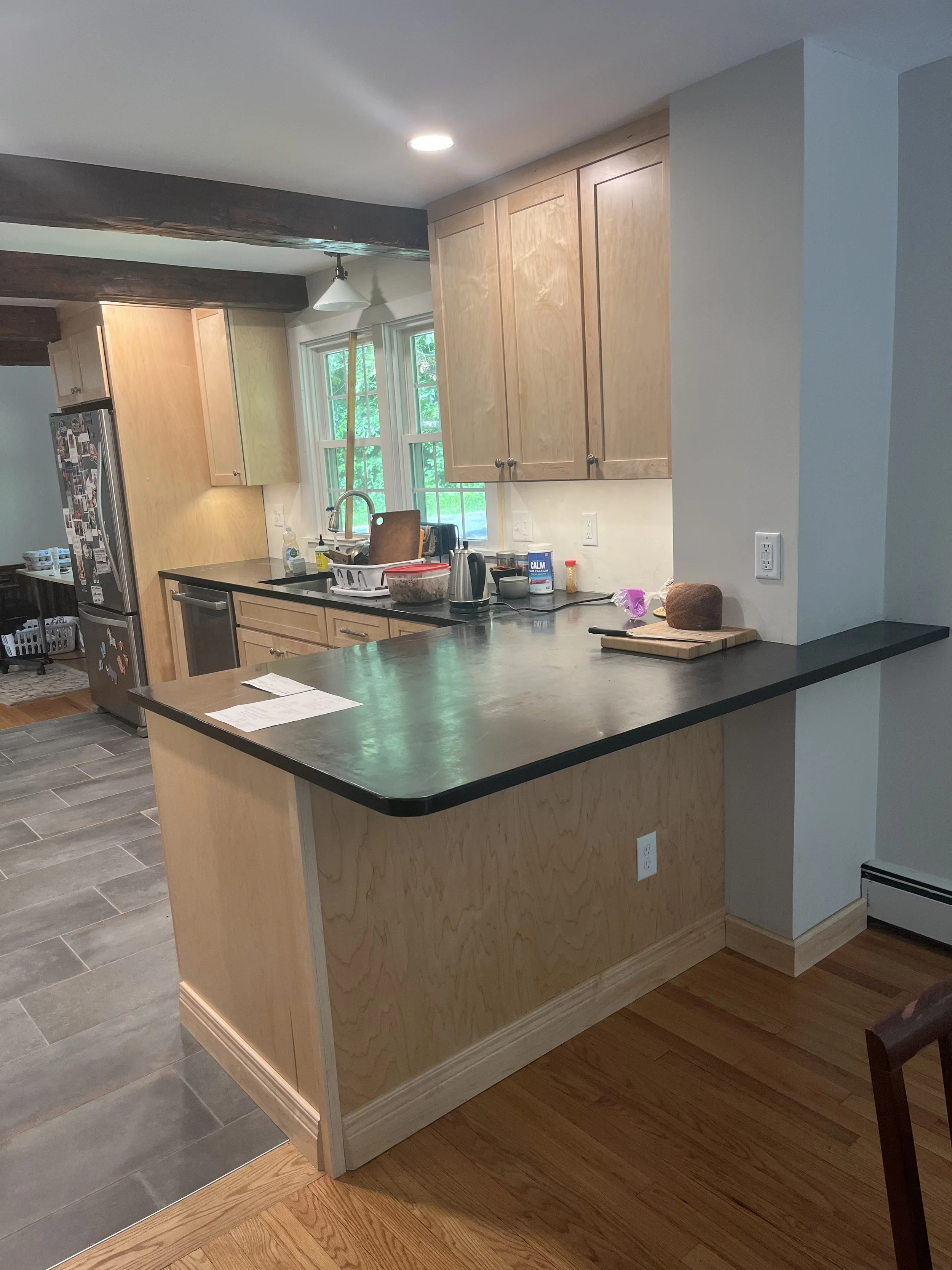 Kitchen with wood cabinets, black countertop, stainless steel appliances, and a window over the sink. Items on the counter include a cutting board, bread, and various kitchen supplies. Floors are a mix of gray tile and wood.
