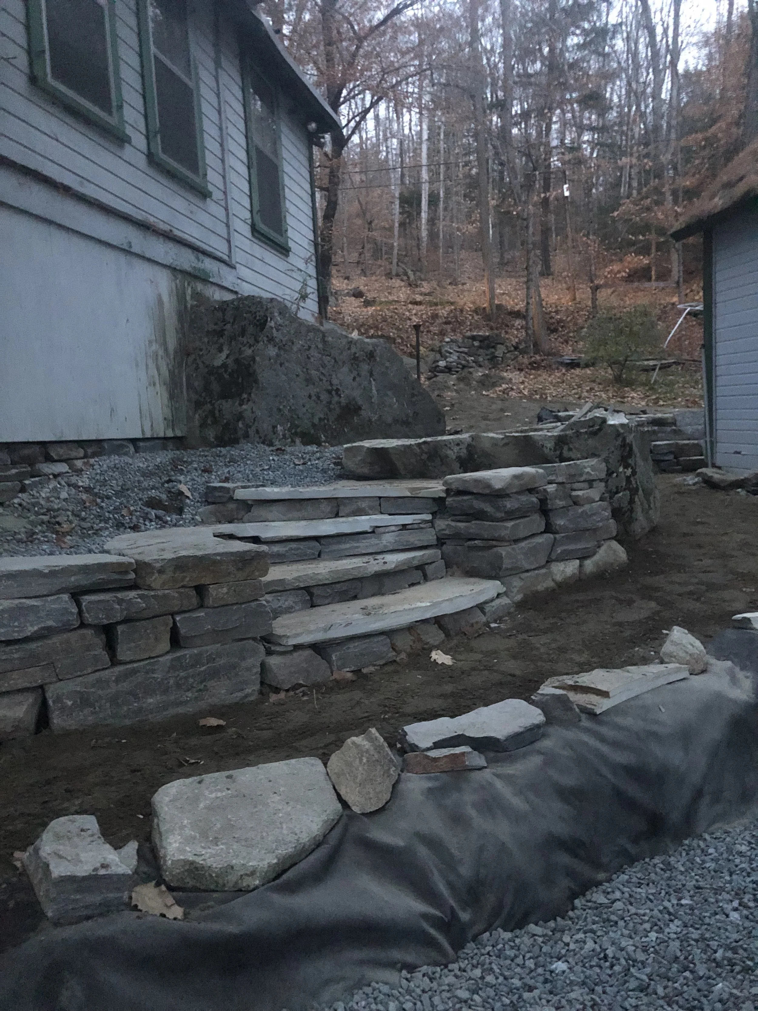 View of a stone staircase leading up to a house, with surrounding rocks and dirt, in a wooded area during autumn.