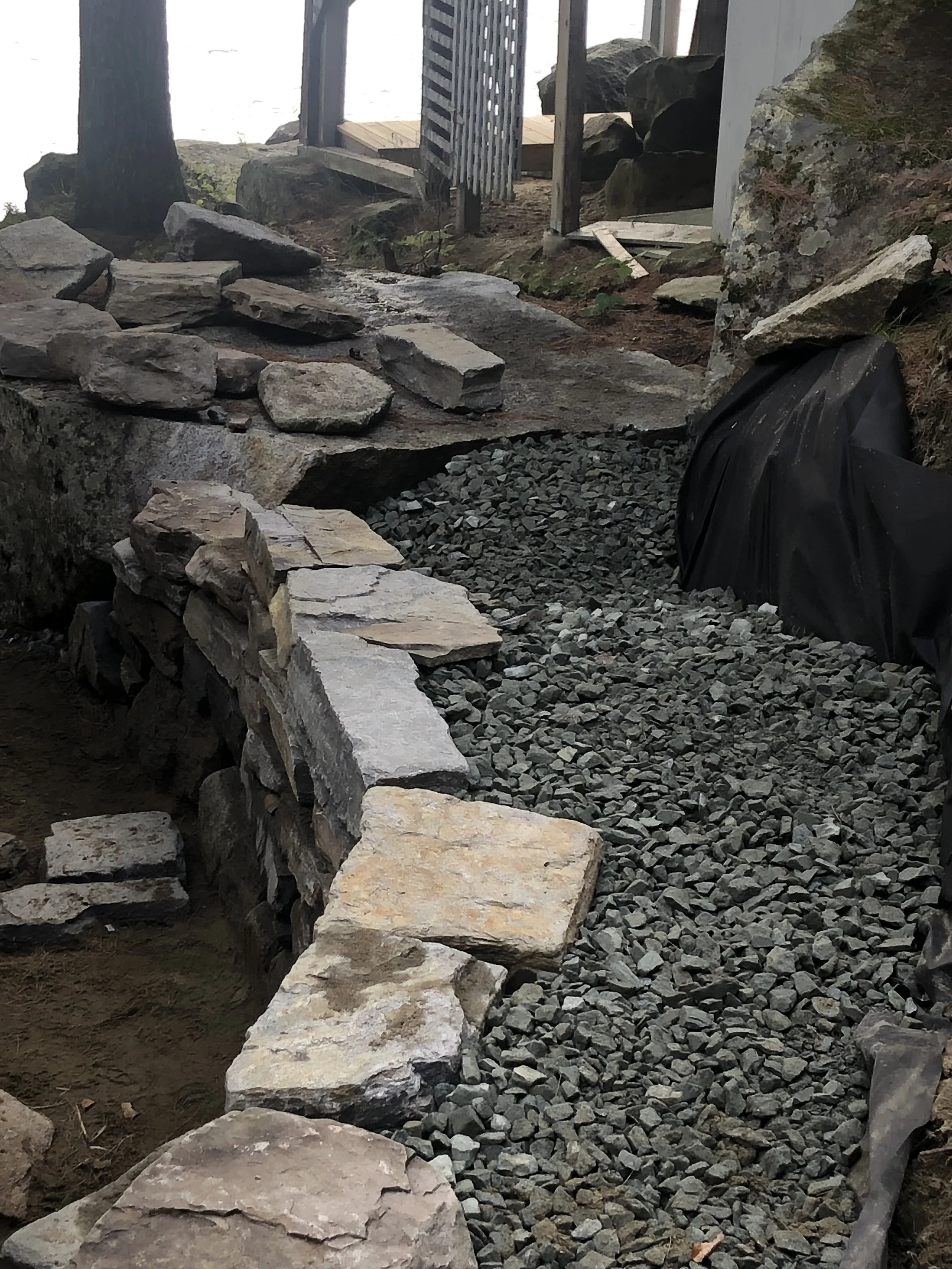 This image shows a construction site with a stone pathway being built. Large stones are arranged along the edge, and smaller gravel fills the section between the stones. There is a black tarp covering a part of the ground on the right. In the backgro