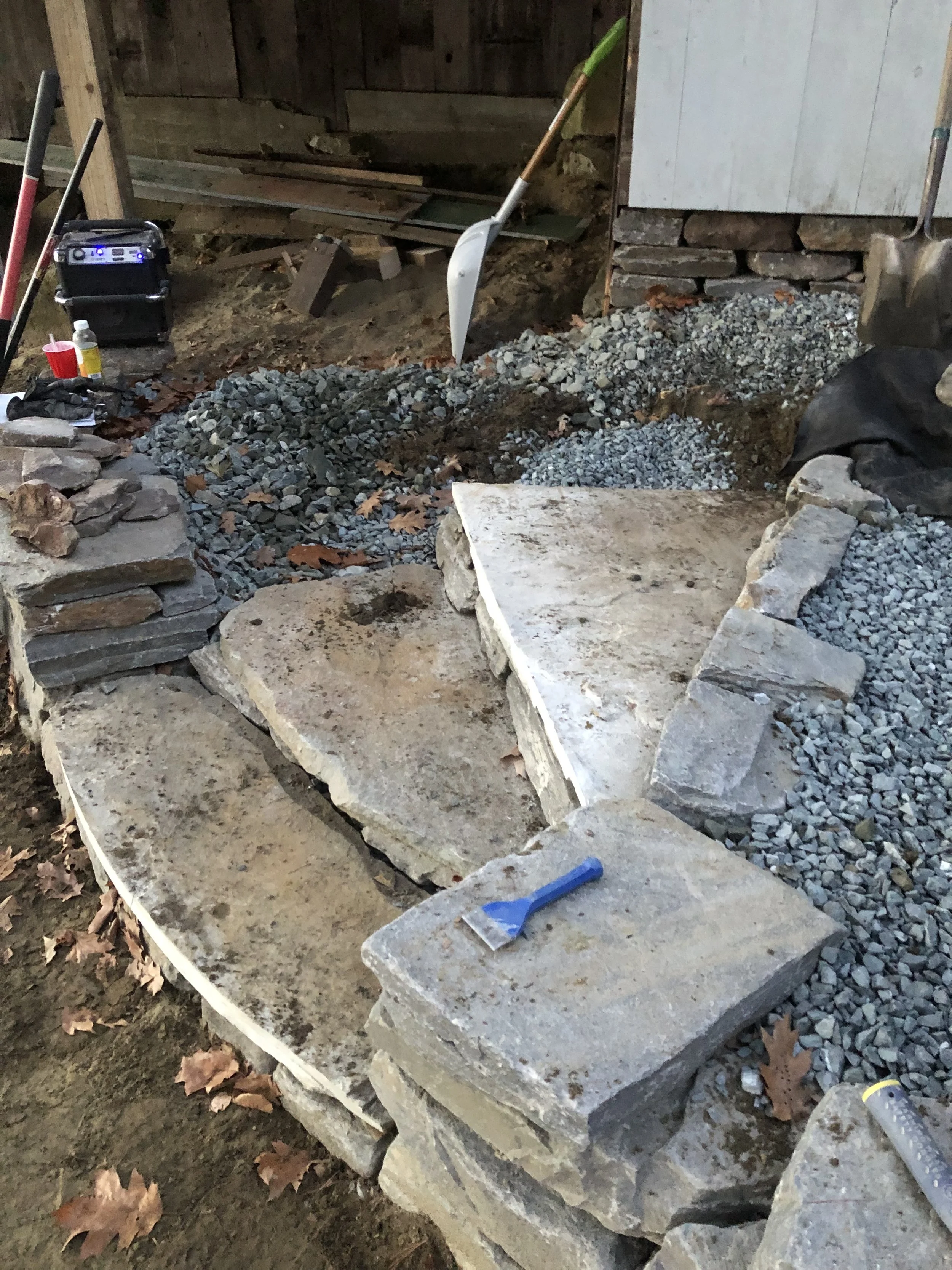 Under construction or renovation site with stone steps, gravel, tools, and equipment for building or repairing a stone pathway or wall.