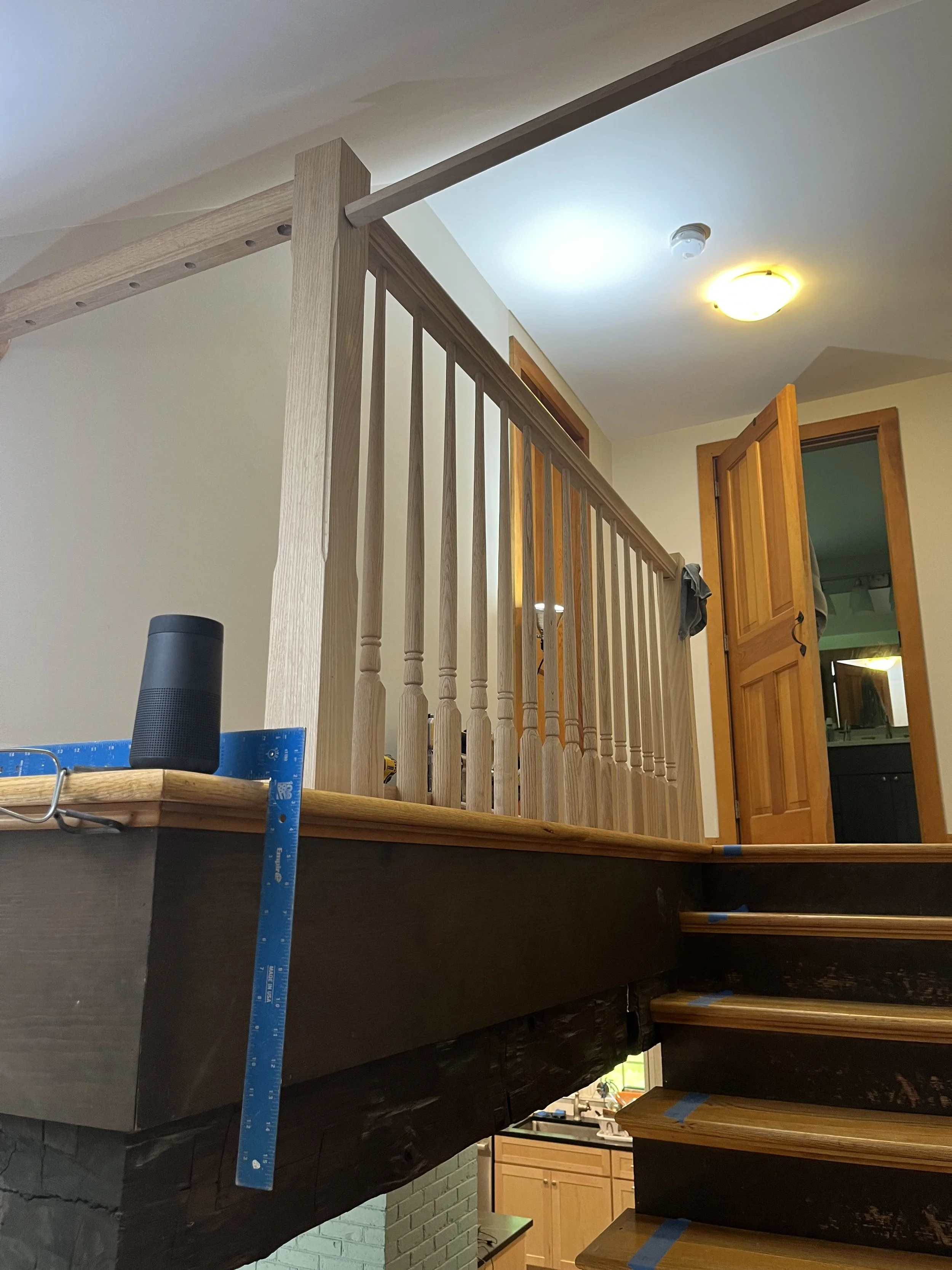 Interior of a house showing stairs leading to a loft or second level, with a wooden railing and open door to another room, ceiling light, and construction materials nearby.
